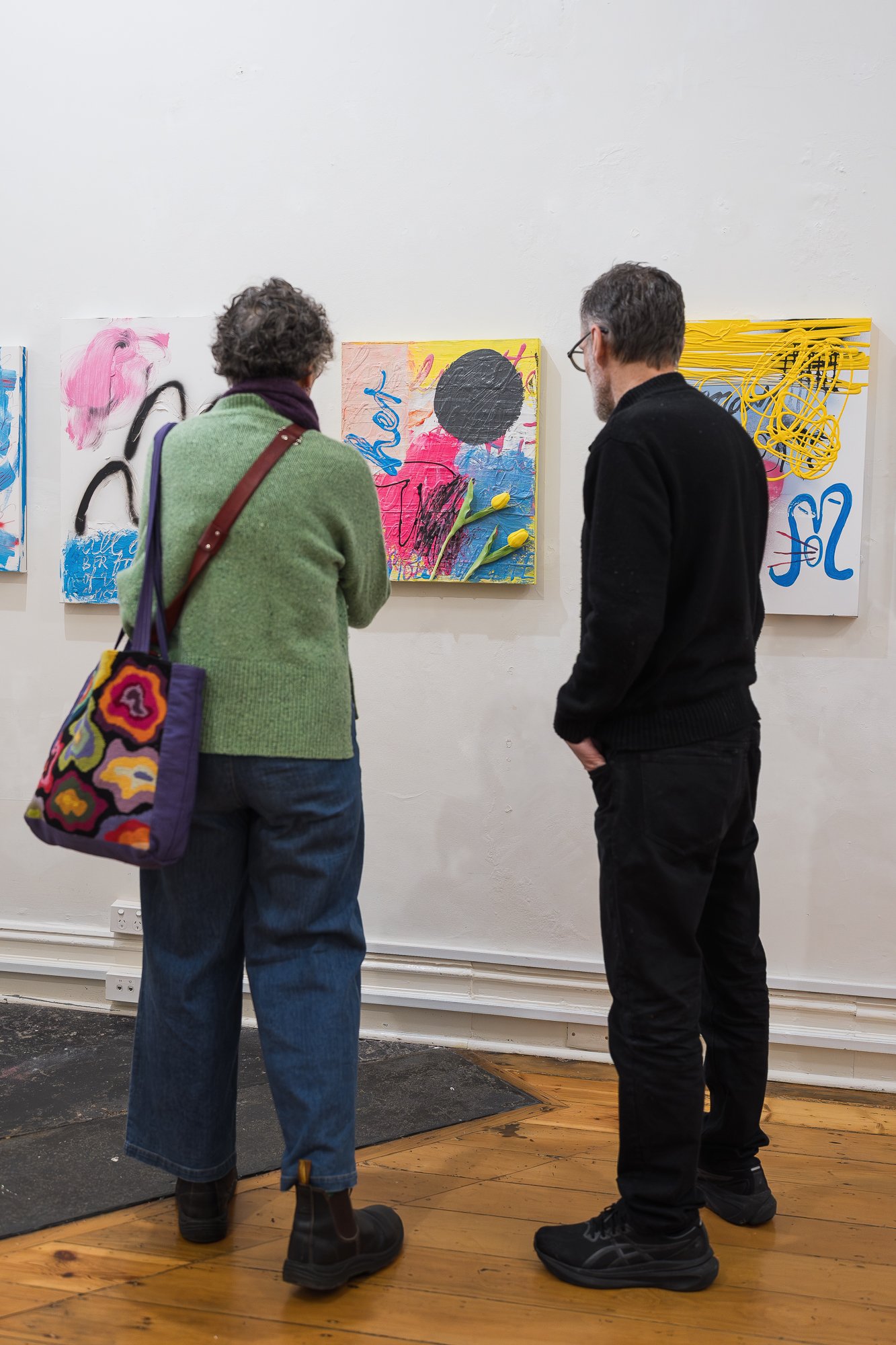 Two visitors viewed from behind, standing in the gallery looking at a wall of colourful abstract paintings.
