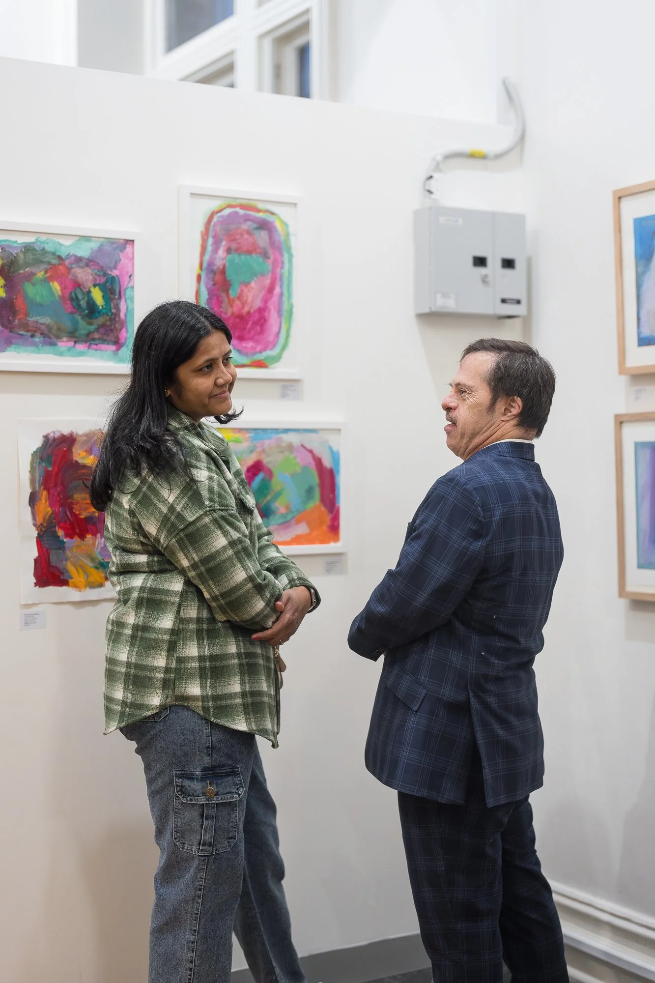 Two people in conversation in front of a wall of vibrant small works — the man in a navy check blazer mid-sentence, both engaged.