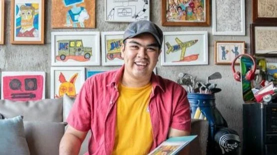 A portrait of a smiling diverse artist, in bright clothing smiling at the camera and holding an art folio with a collection of coloured artworks hanging on the wall ion the background.