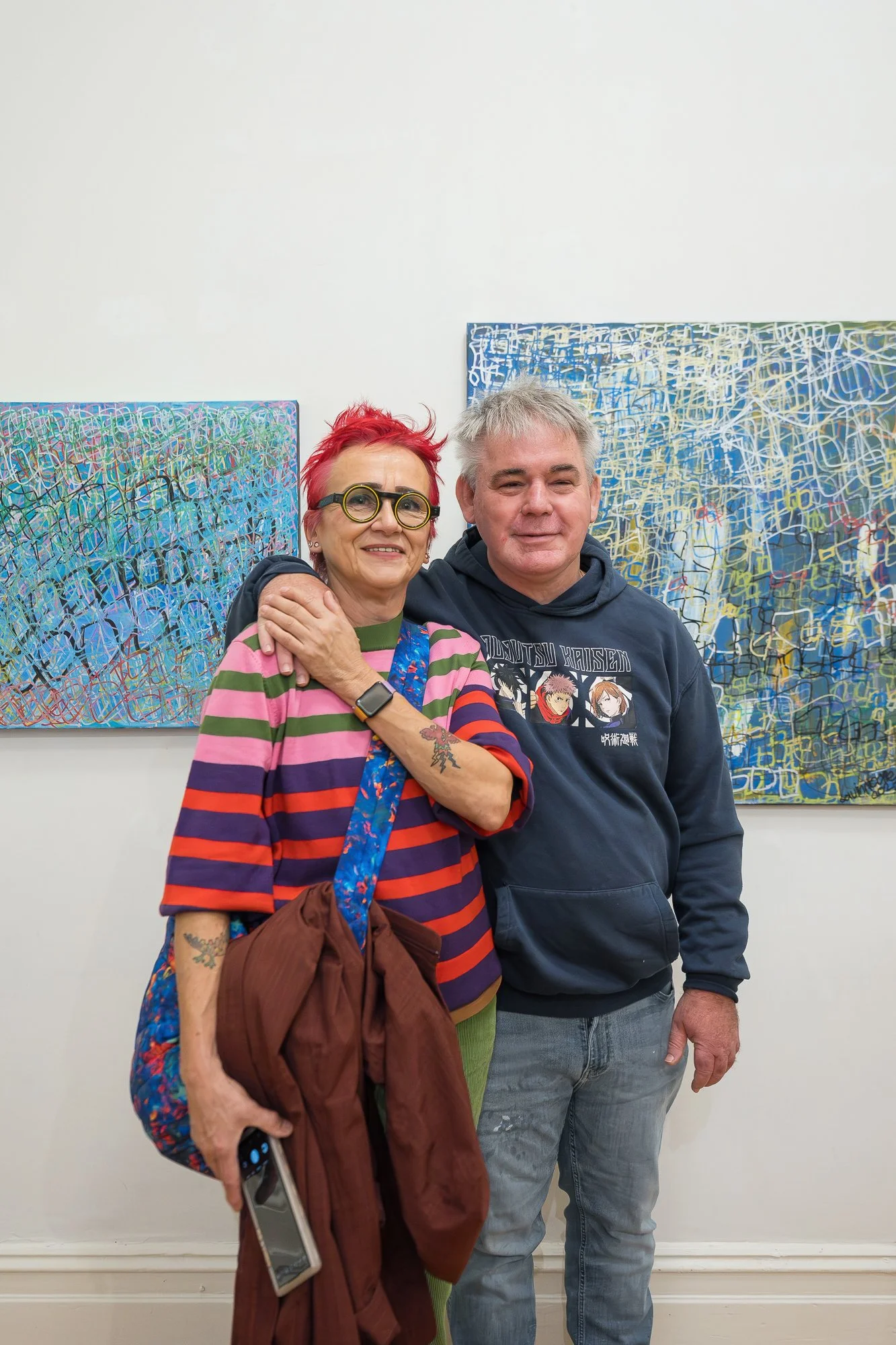 Two people smile warmly together in front of two large densely layered abstract paintings in blue and yellow-green.