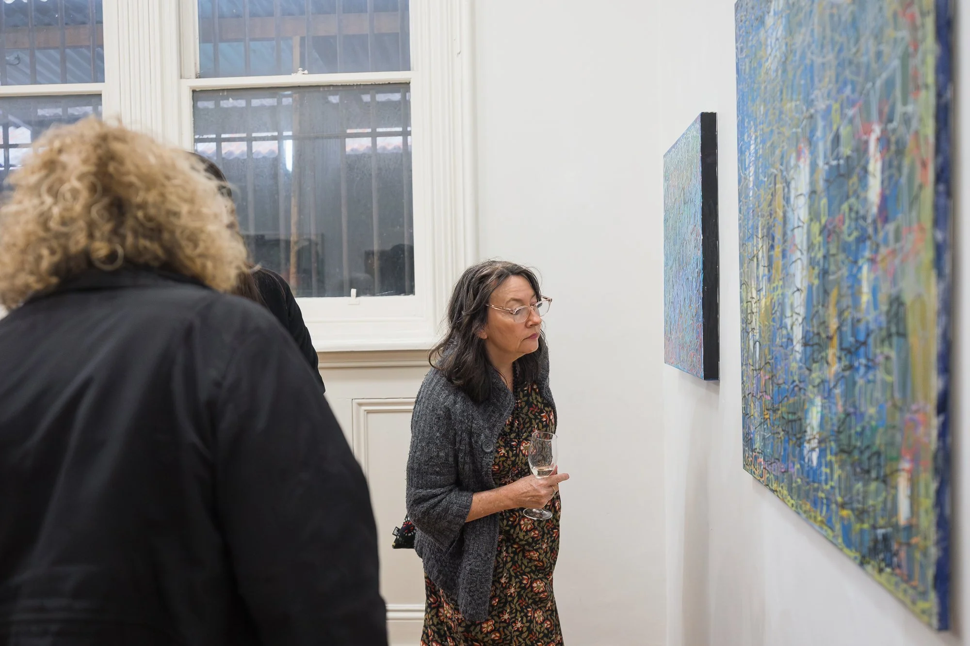 A woman holds a glass of wine as she quietly studies two densely layered abstract paintings in blue and green.