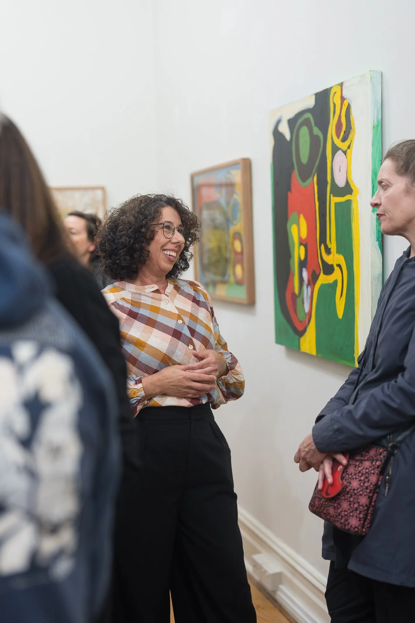 Two women laugh together in conversation, with a large bold abstract painting in greens, reds and yellows on the wall behind them.