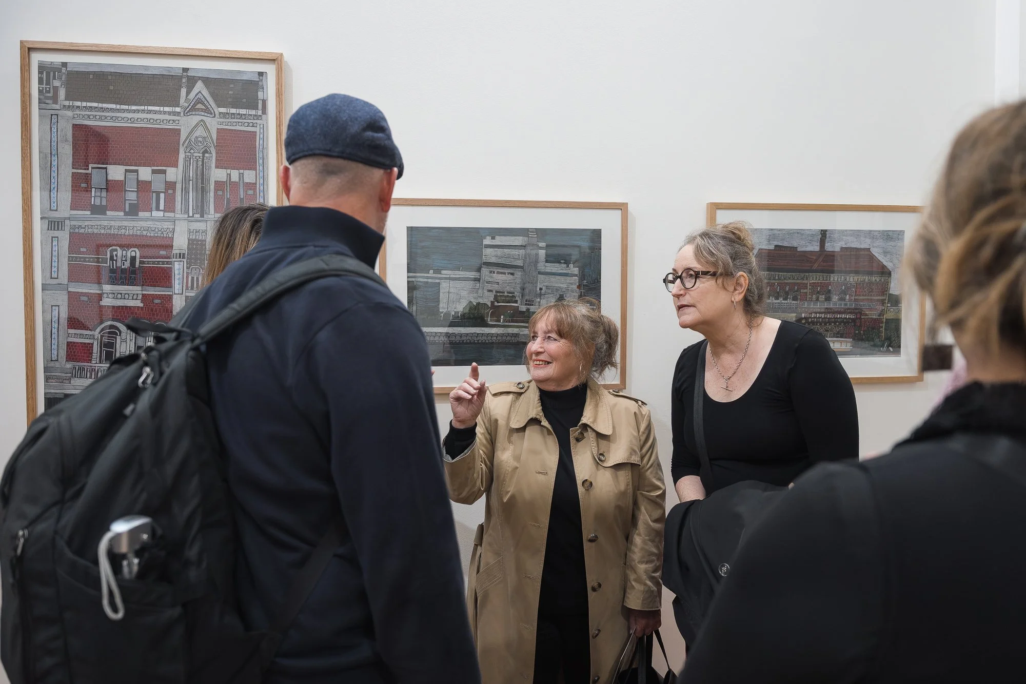 Group of visitors in dark clothing in conversation in front of a row of framed architectural illustrations of Melbourne buildings.