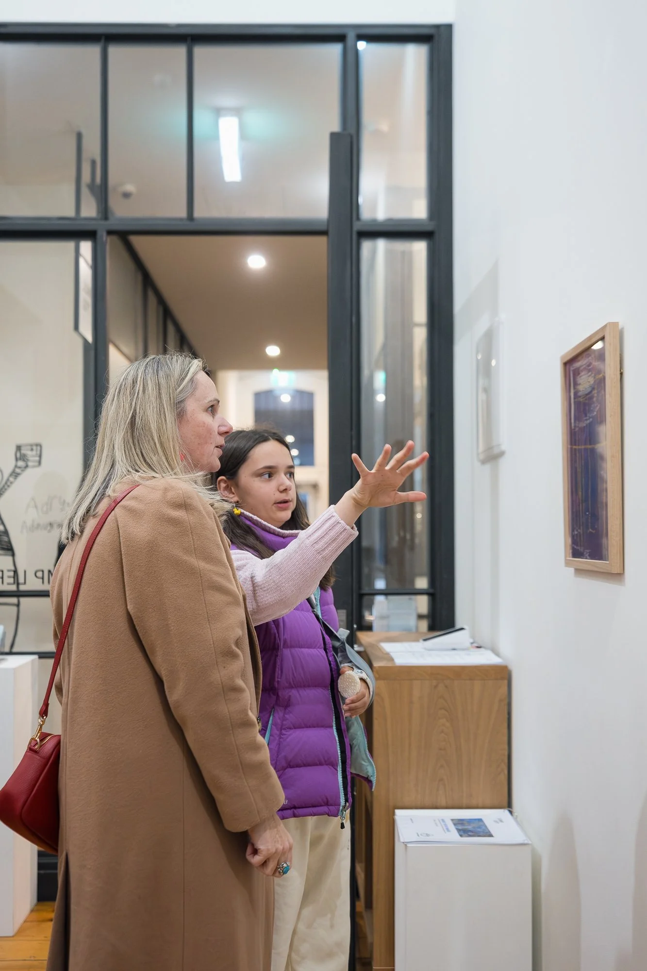 A young person in a purple vest gestures toward a small framed work, sharing their thoughts with the person beside them.