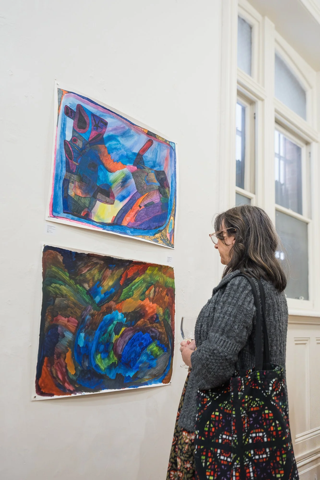 A woman with a glass of wine pauses to take in two vivid works on paper — one a dense blue and purple composition, the other dark and richly coloured.
