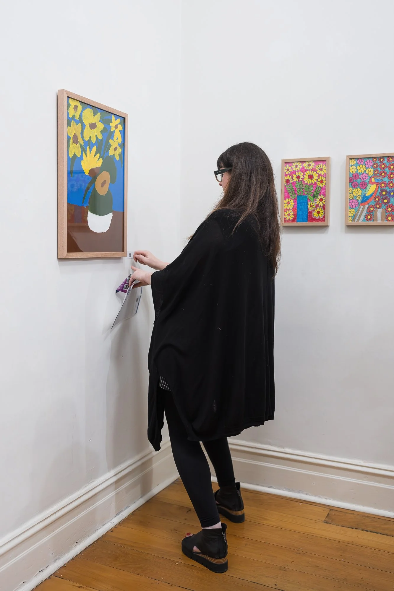 Woman in black viewed from behind, adjusting a tag/label beside a framed sunflower still-life painting, with two floral paintings visible on the right-hand wall.