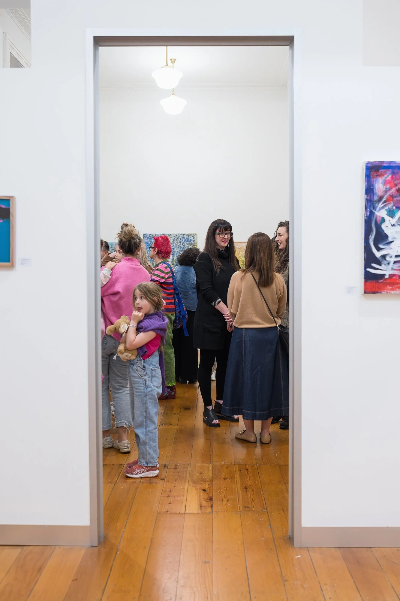 A busy gallery room seen through a doorway, packed with guests of all ages, including a young child clutching a stuffed toy.