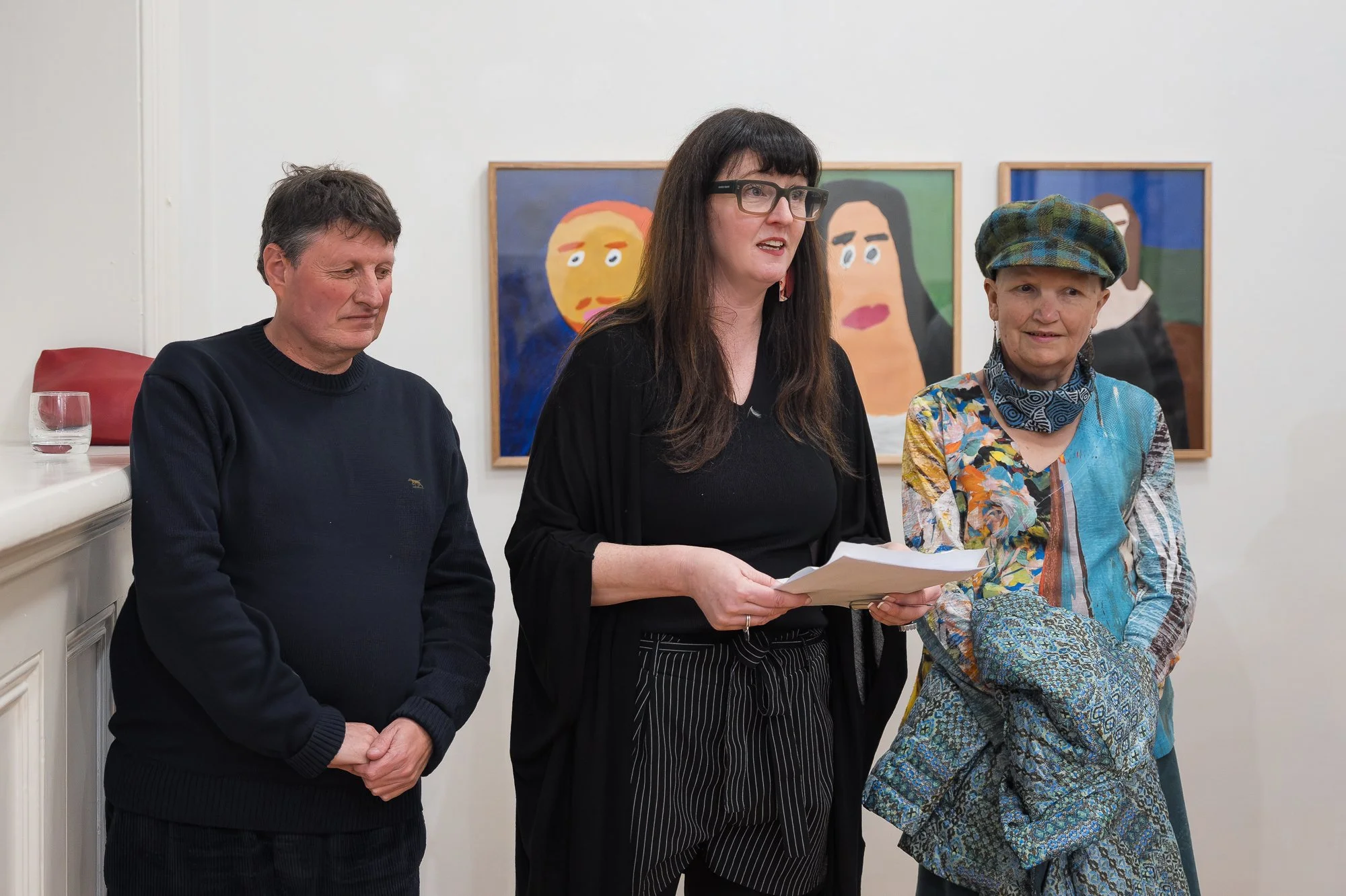 Three people at a small gathering: a man listening, a woman speaking while holding papers, and a woman in a colourful jacket and cap beside her, portrait paintings on the wall behind.