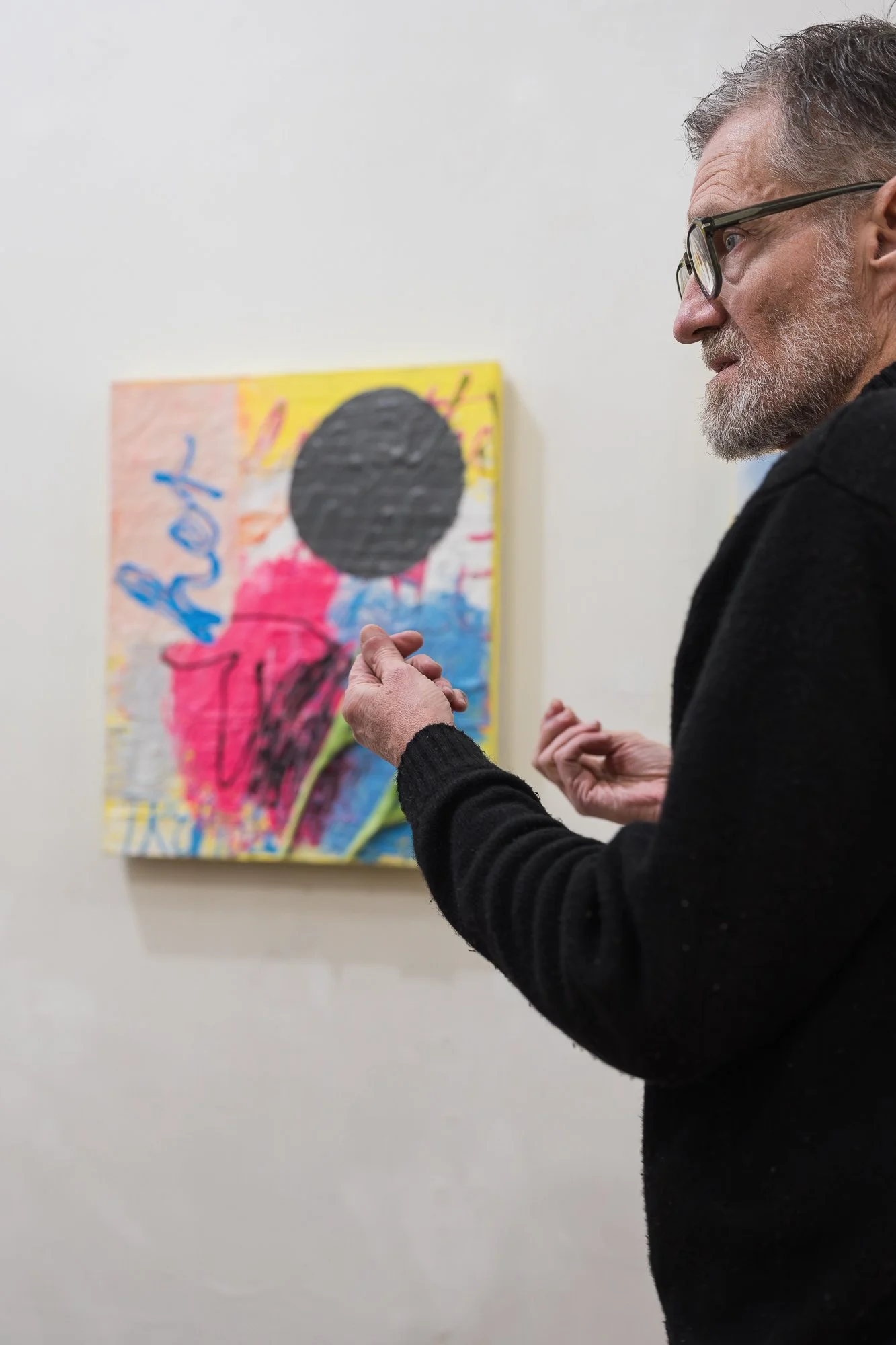 Close-up profile of a bearded man gesturing while speaking, with an abstract mixed-media painting (yellow, pink, black circle motif) on the wall behind him.