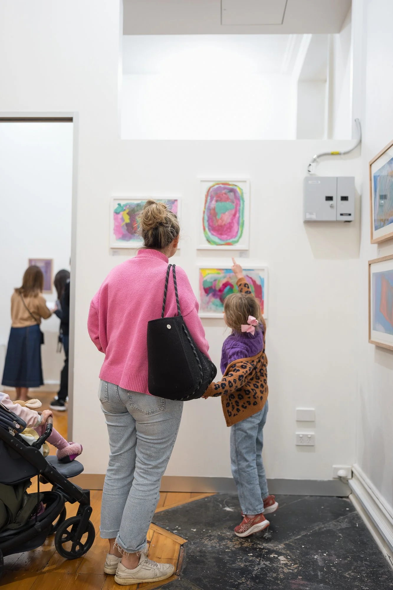 A woman in pink holds a small child's hand as the child points excitedly at a bright, colourful work on the wall.