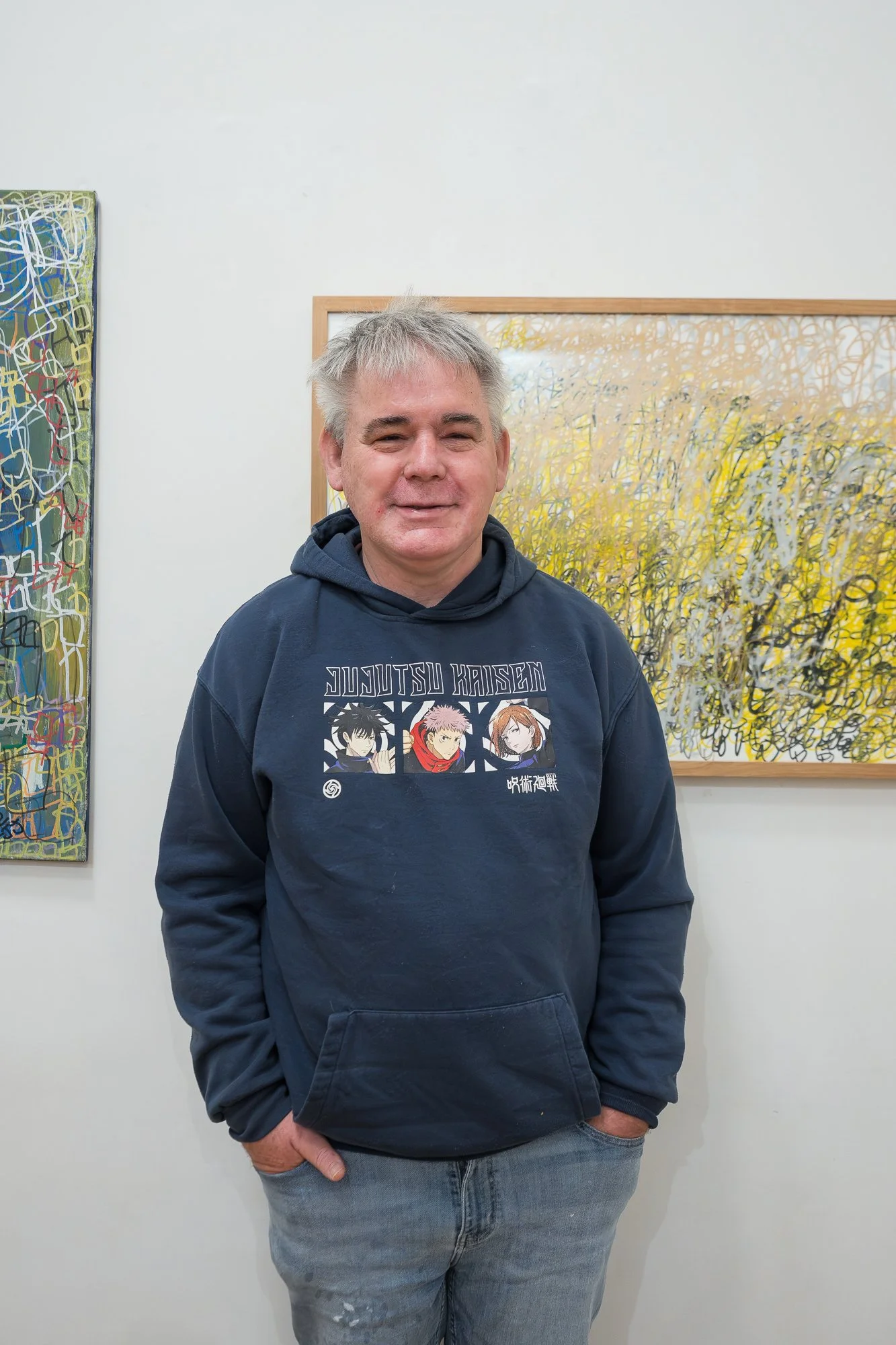 A man in a navy hoodie smiles warmly in front of two densely layered abstract works in yellow, green and blue.