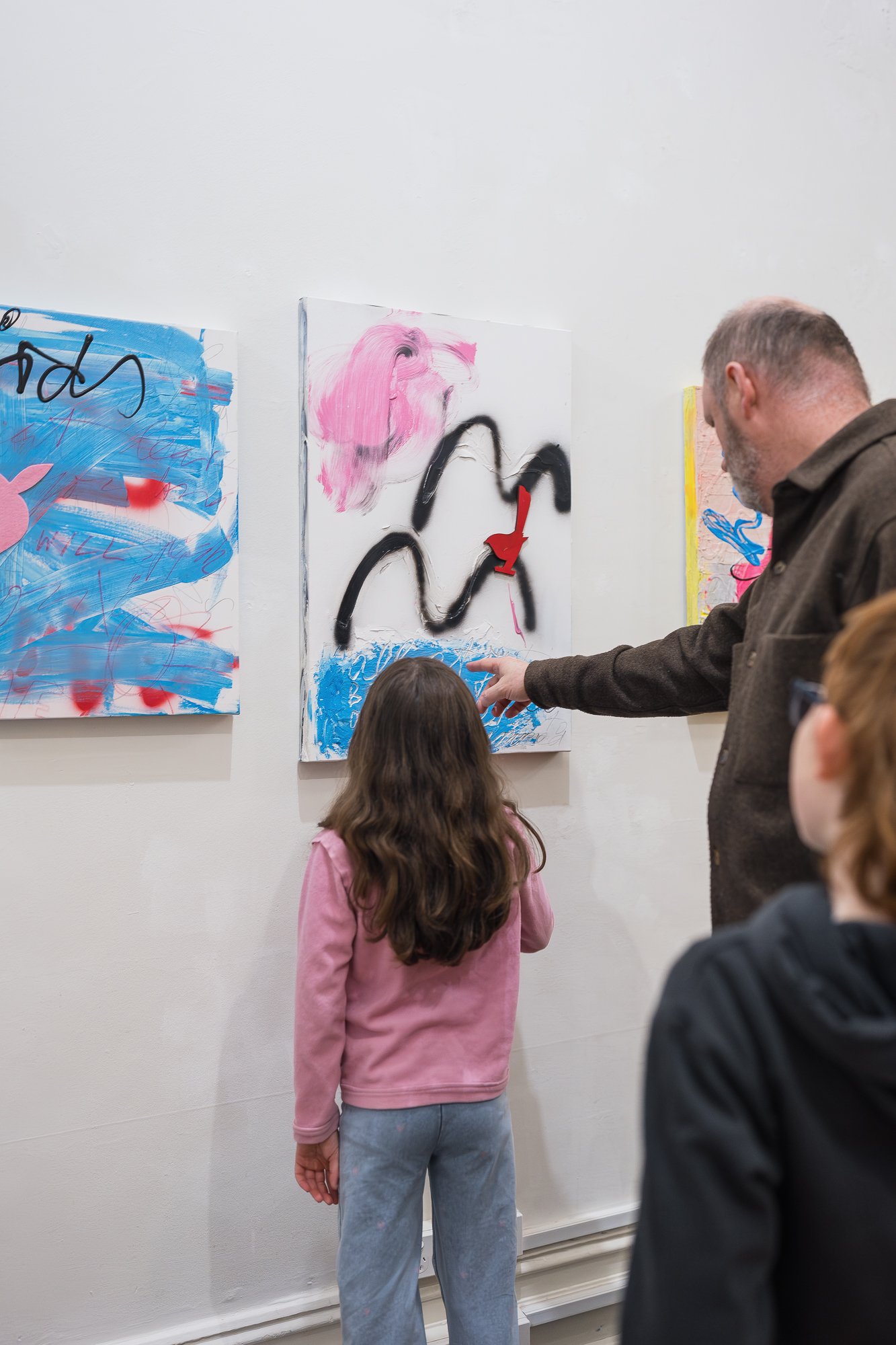 Close-up of a man and child viewing abstract mixed-media paintings (blue, pink and black gestural marks with a red bird cut-out) on a gallery wall, man pointing to a detail.