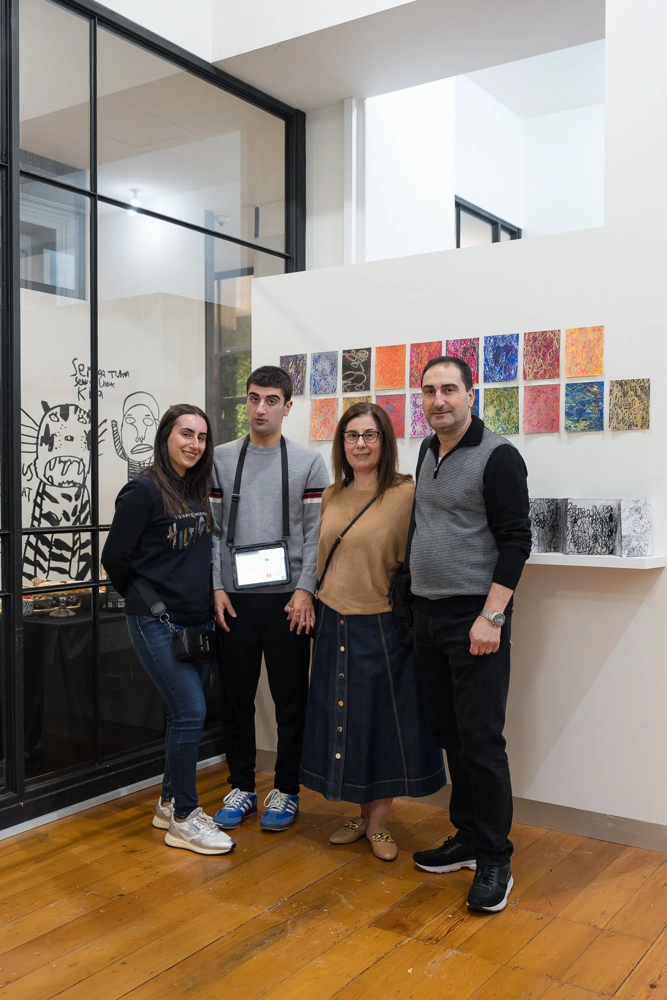A family of four smile together in the gallery, a vivid grid of small colourful works displayed behind them.