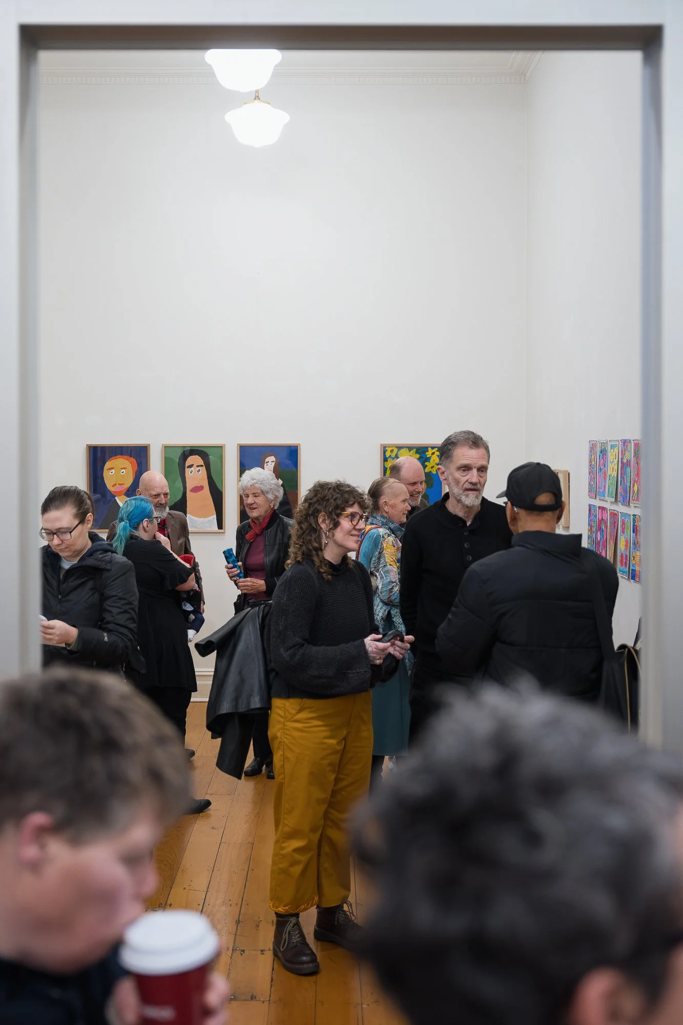 Wide shot through a doorway of a crowded gallery room, visitors gathered near portrait and floral artworks.