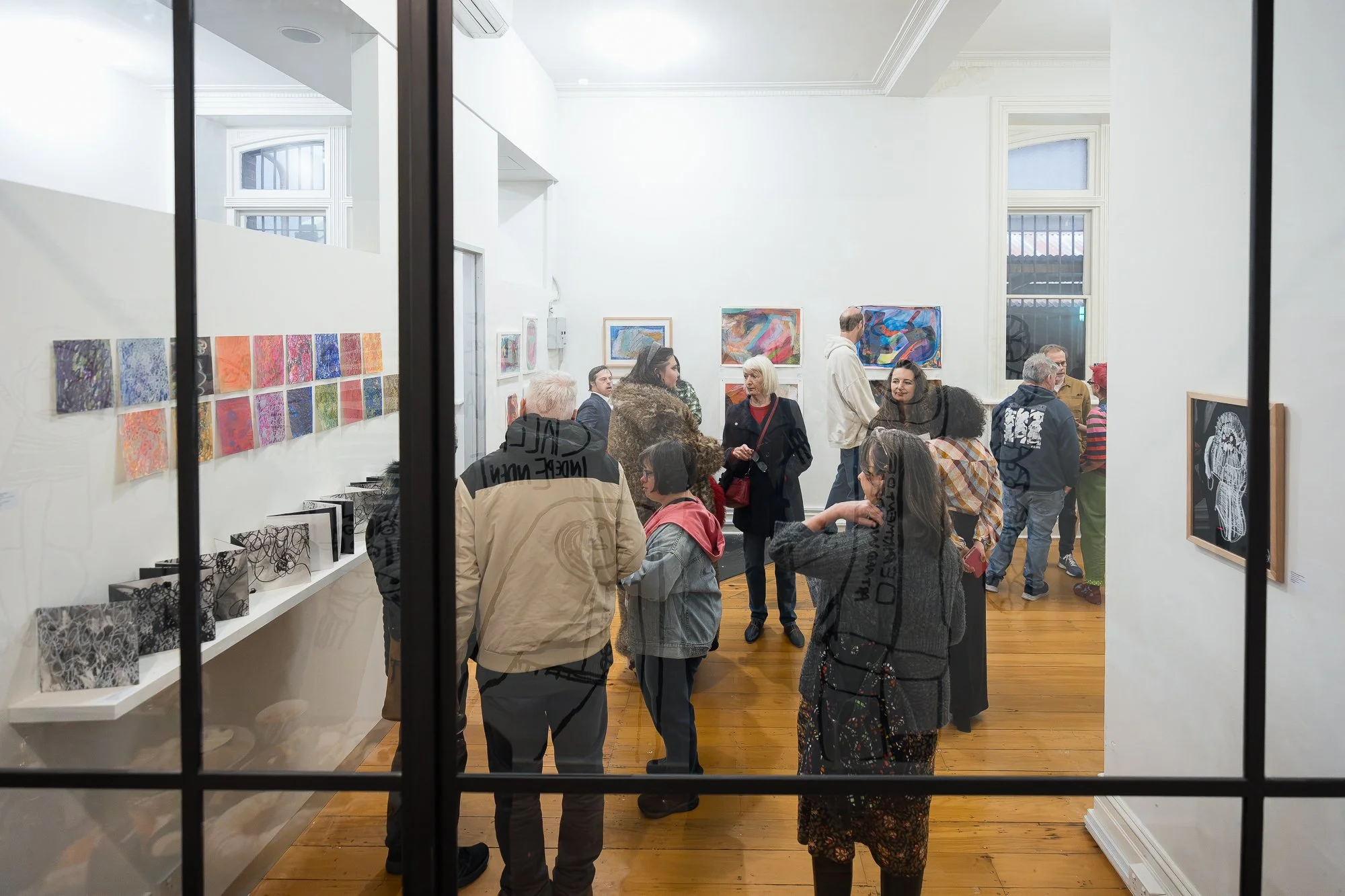 The gallery viewed through the glass partition — a buzzing crowd moving through the space, colourful works covering every wall.