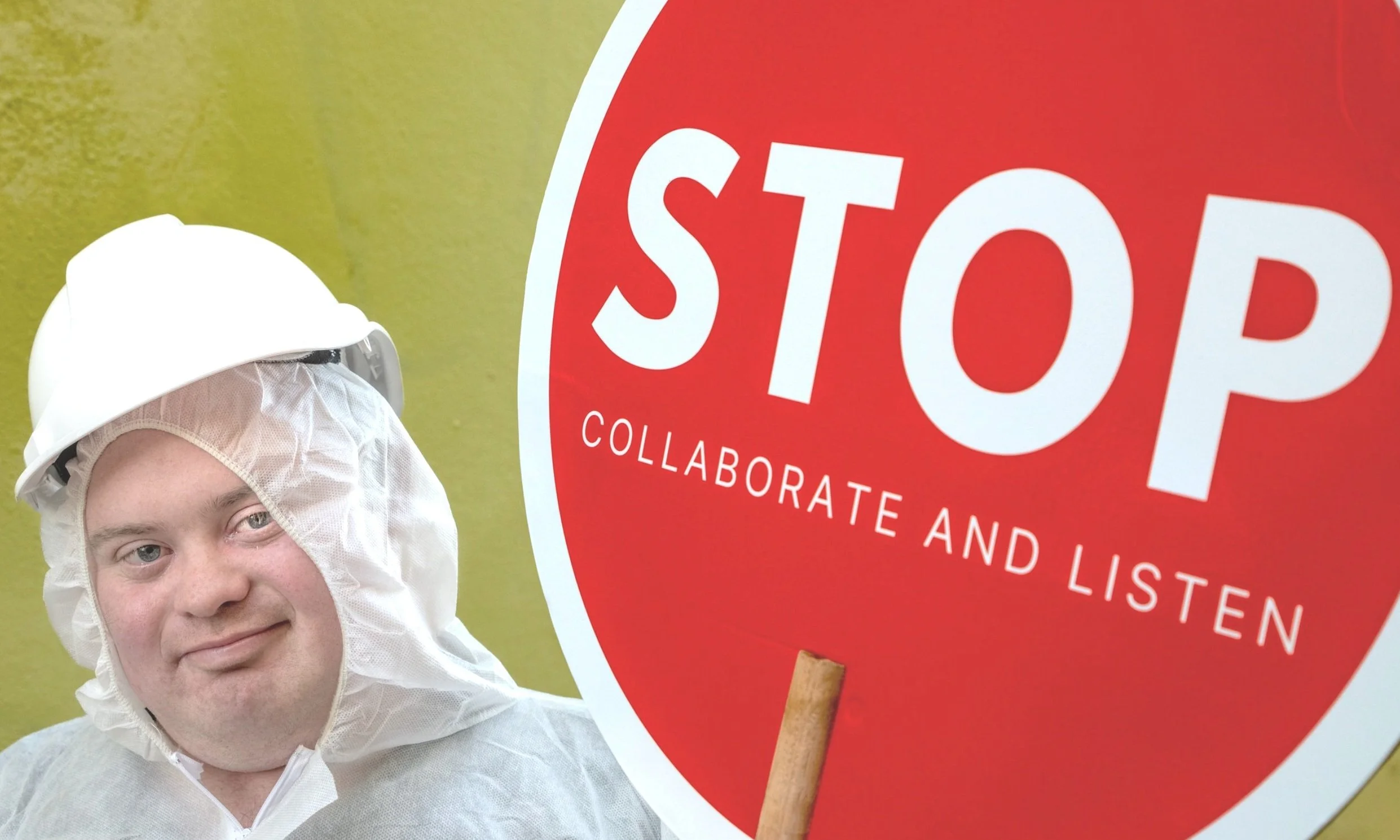 A diverse male in a rain jacket with a hard-hat, holding a red sign that reads 'Stop, collaborate and listen'.