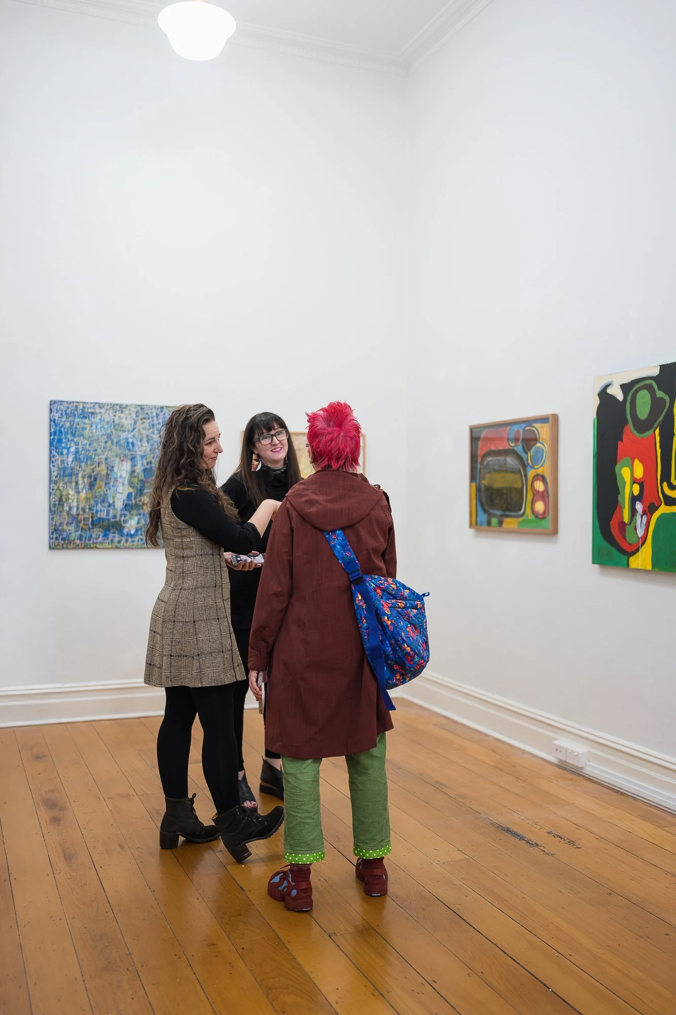 Three people chat in a bright gallery space surrounded by large colourful paintings.