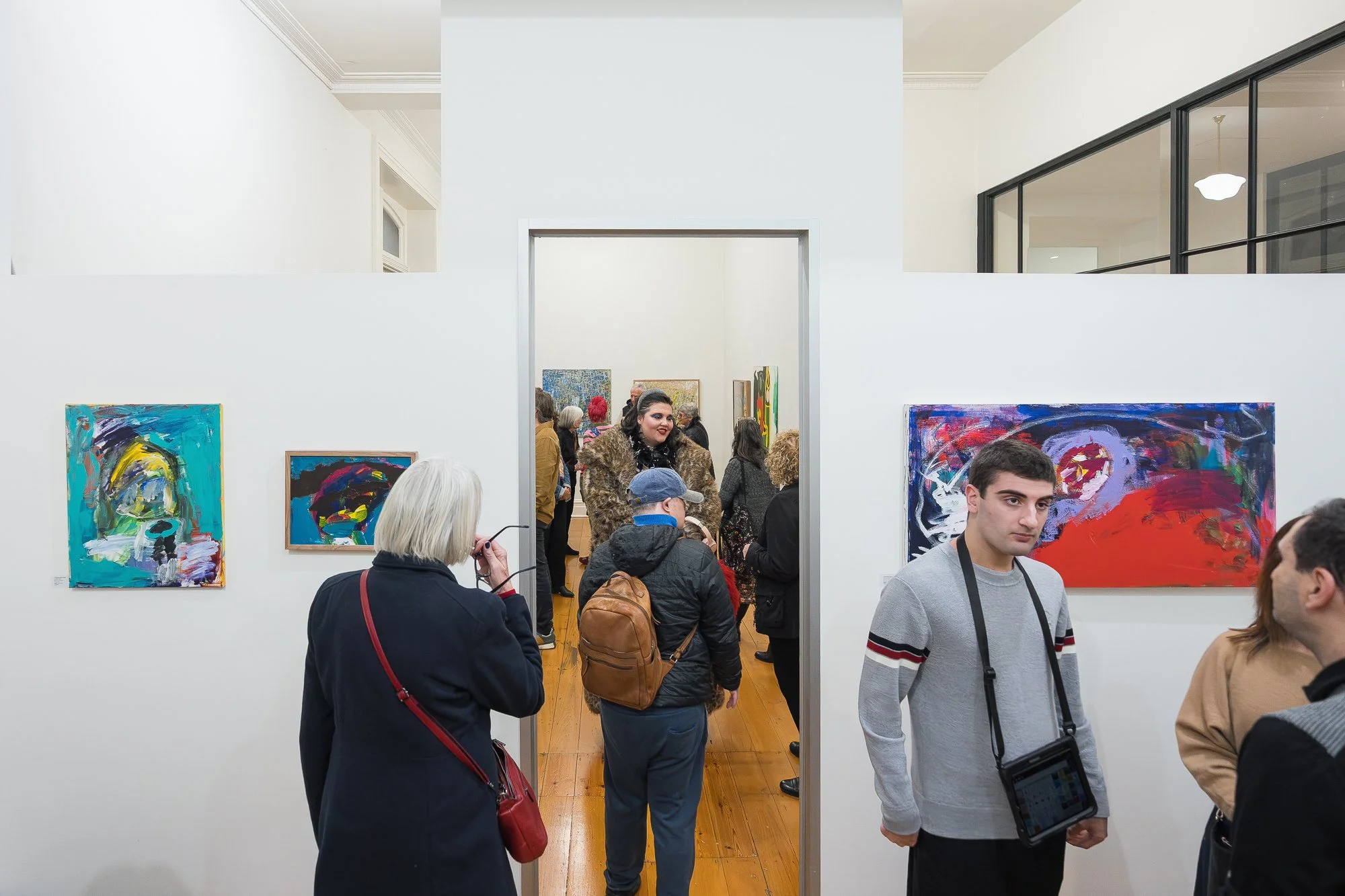 Guests flow between gallery rooms, pausing at paintings on every wall — a full, lively opening night crowd.