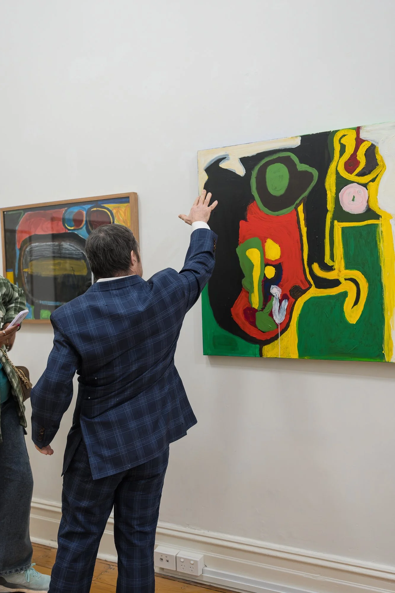 A man in a navy check blazer reaches up to gesture at a large bold abstract painting in greens, reds and yellows, explaining the work to a visitor beside him.