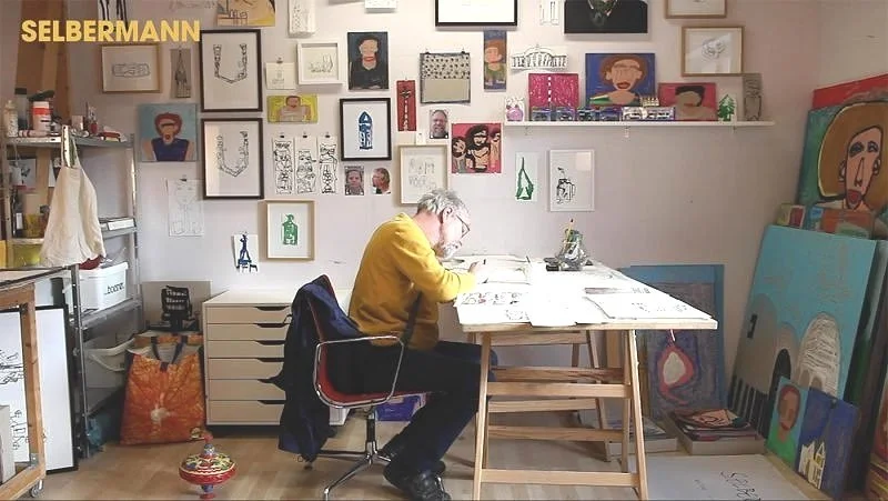 A view of an artist's studio with an artist working at the desk, surrounded by a vast collection of artworks, materials and tools.