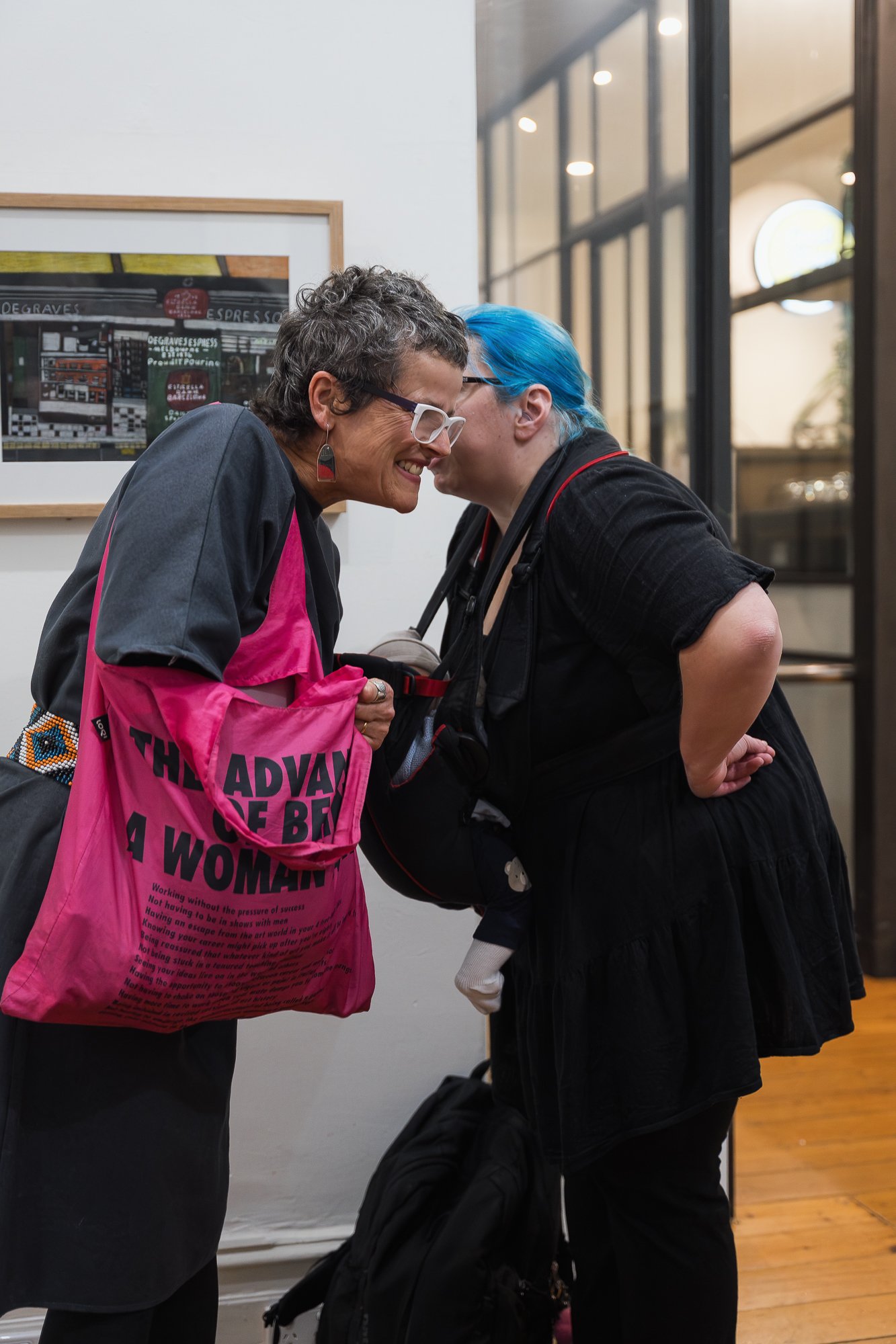 Two women smiling closely face-to-face, one carrying a pink tote bag printed with "The Advantages of Being a Woman" text.