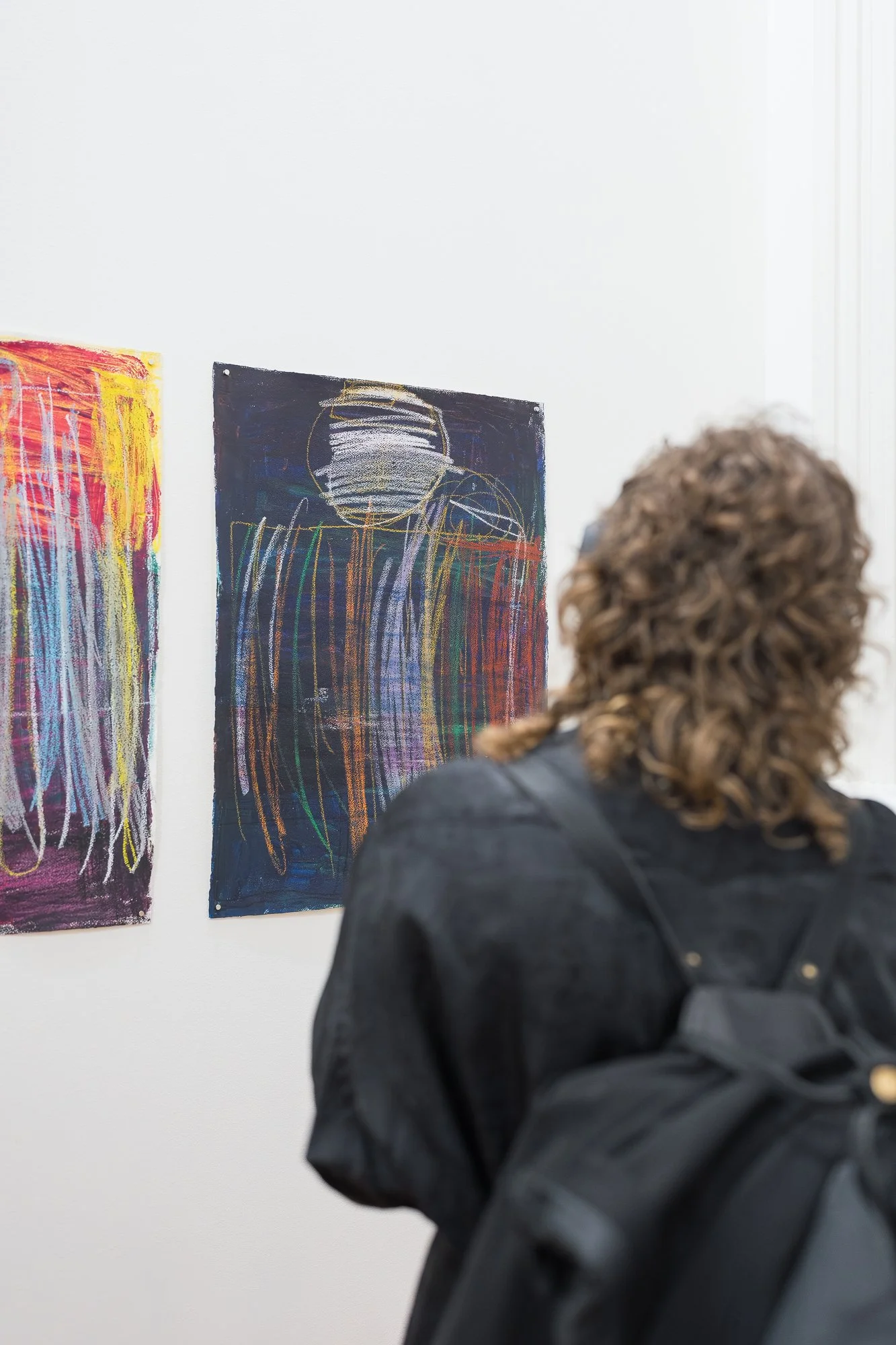 A visitor with curly hair looks closely at two expressive works on paper — one fiery with reds and yellows, the other dark with layered coloured lines and a circular form.
