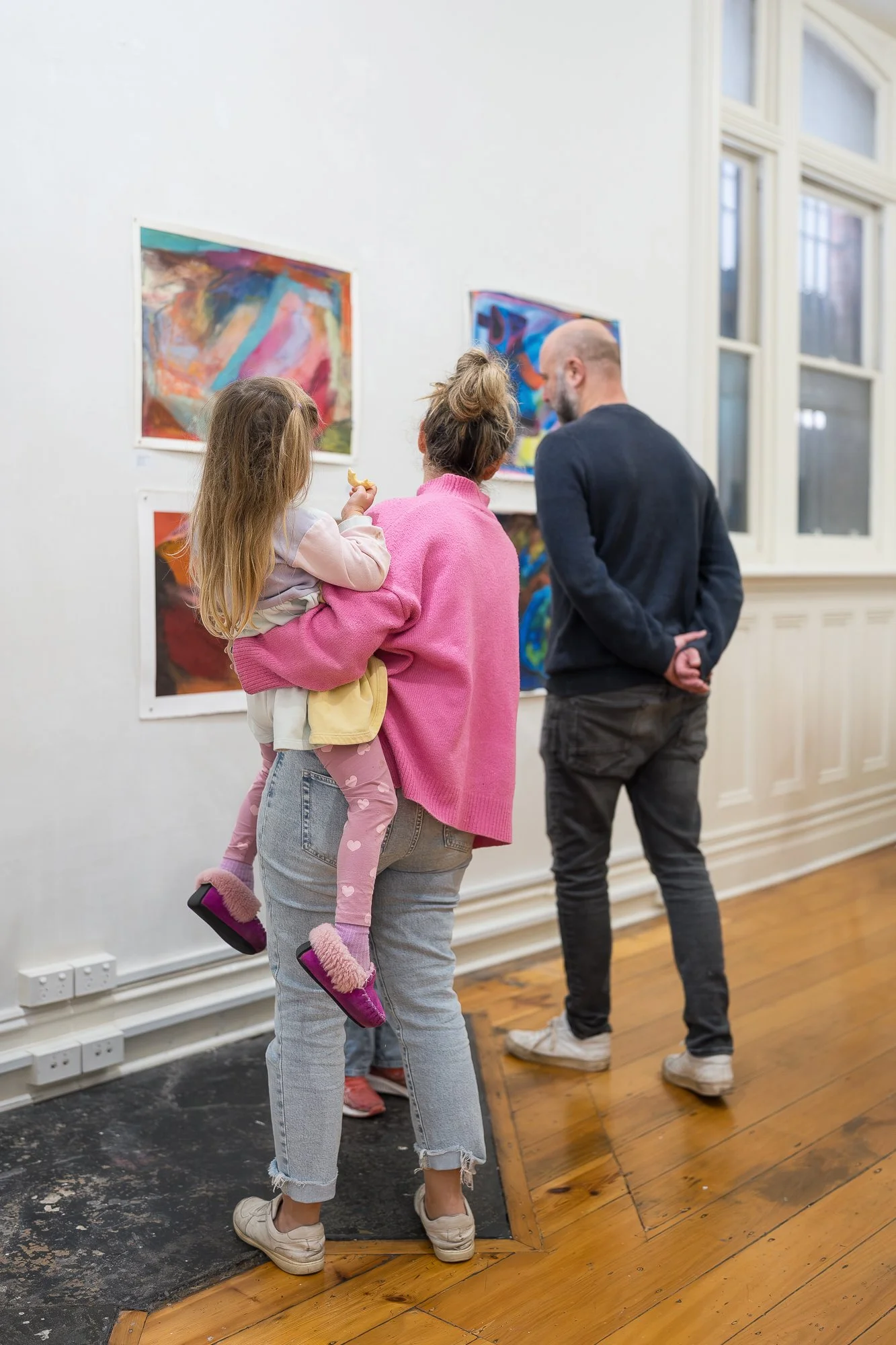 A woman in pink holds a small child on her hip as they look closely at colourful abstract works alongside a man.