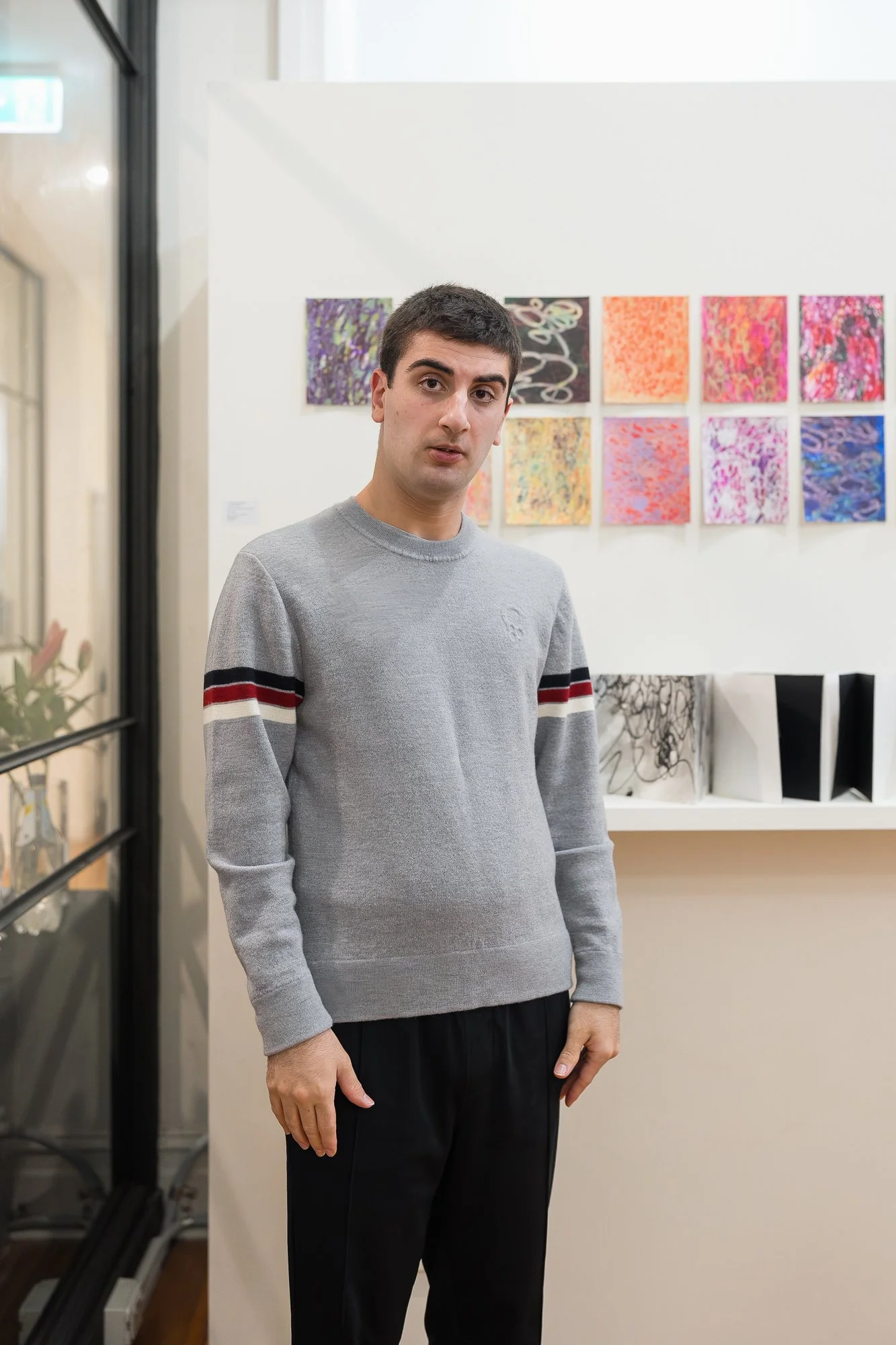A young man in a grey sweater stands in front of a grid of small, vibrant abstract works on the gallery wall.