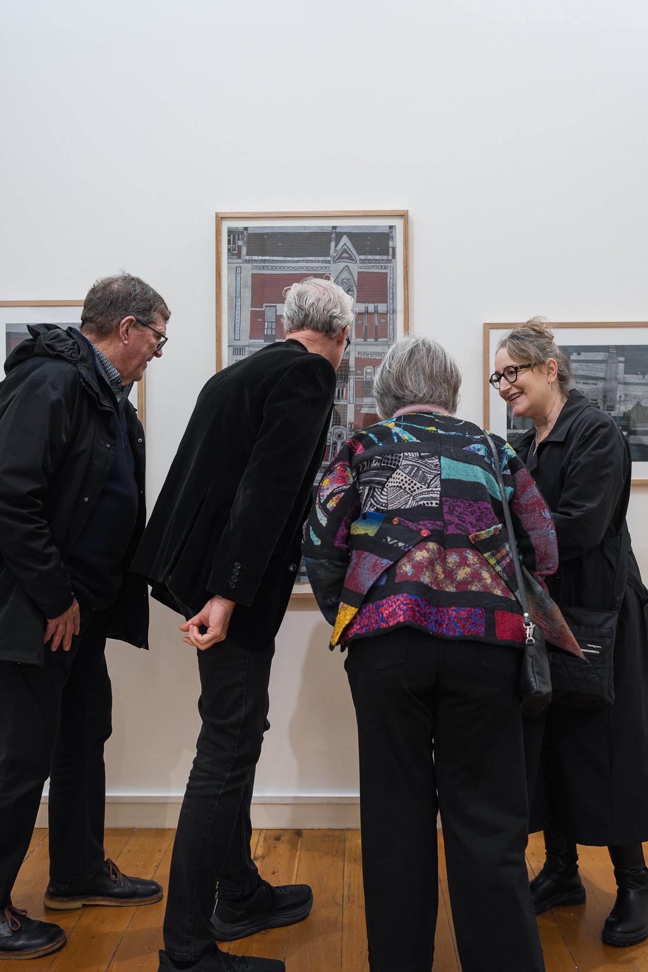Two men and two women in dark clothing gathered closely to view a framed architectural illustration of a heritage building facade on the gallery wall.