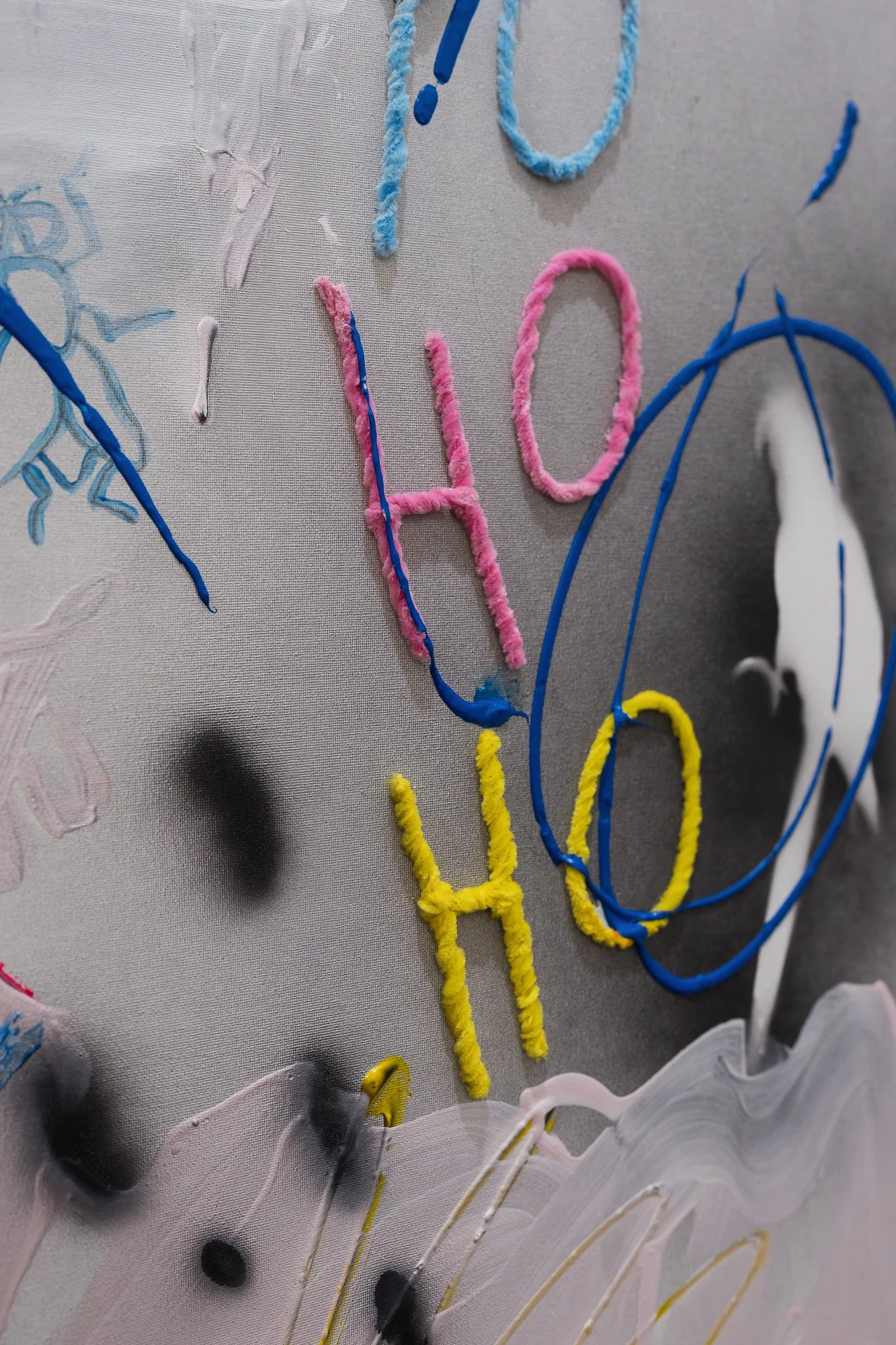  Close-up detail of a mixed-media painting with pink, yellow and blue pipe-cleaner text ("HOHO") and a spray-painted bird stencil on grey canvas.
