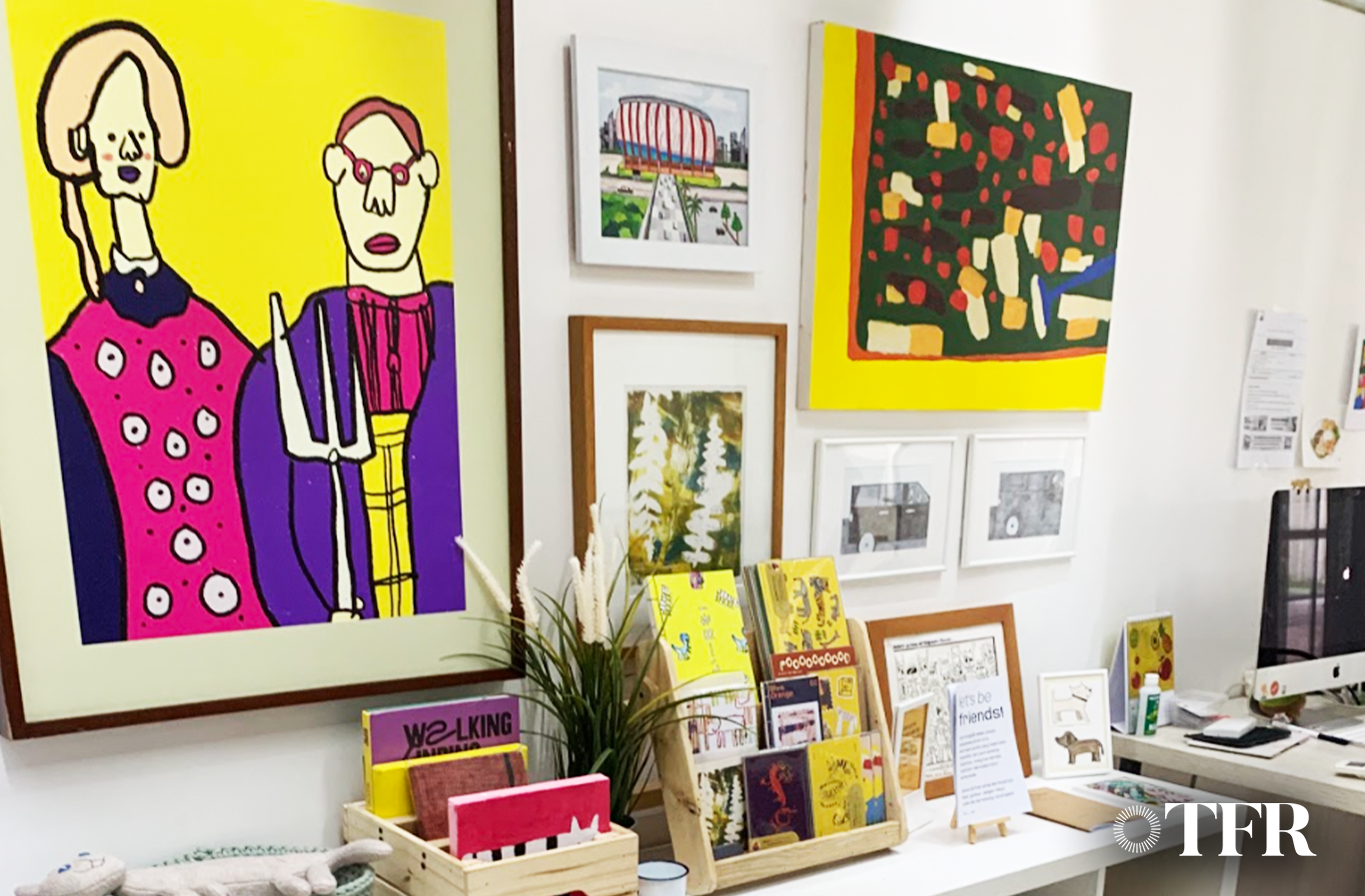Art gallery wall with colorful paintings and prints, including abstract and portrait designs, a wooden display shelf with small booklets, a potted plant, and a computer setup.