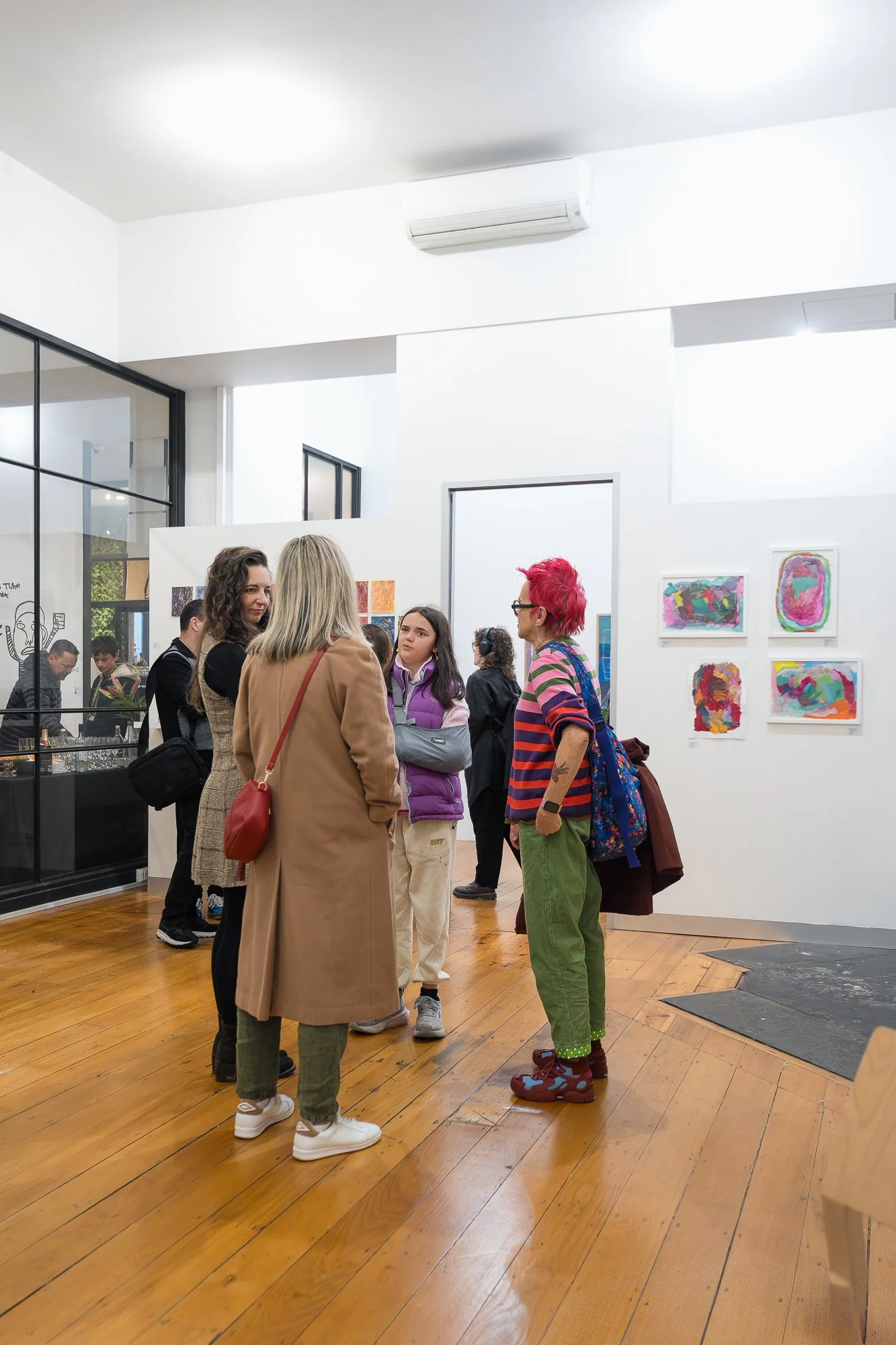 A small group of guests talk in the main gallery space, with a grid of bright, small-scale works visible on the wall behind them.