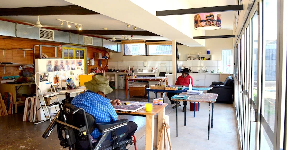 View of a light filled studio, with two artists working on artwork at seperate tables, in front of a wall of windows.