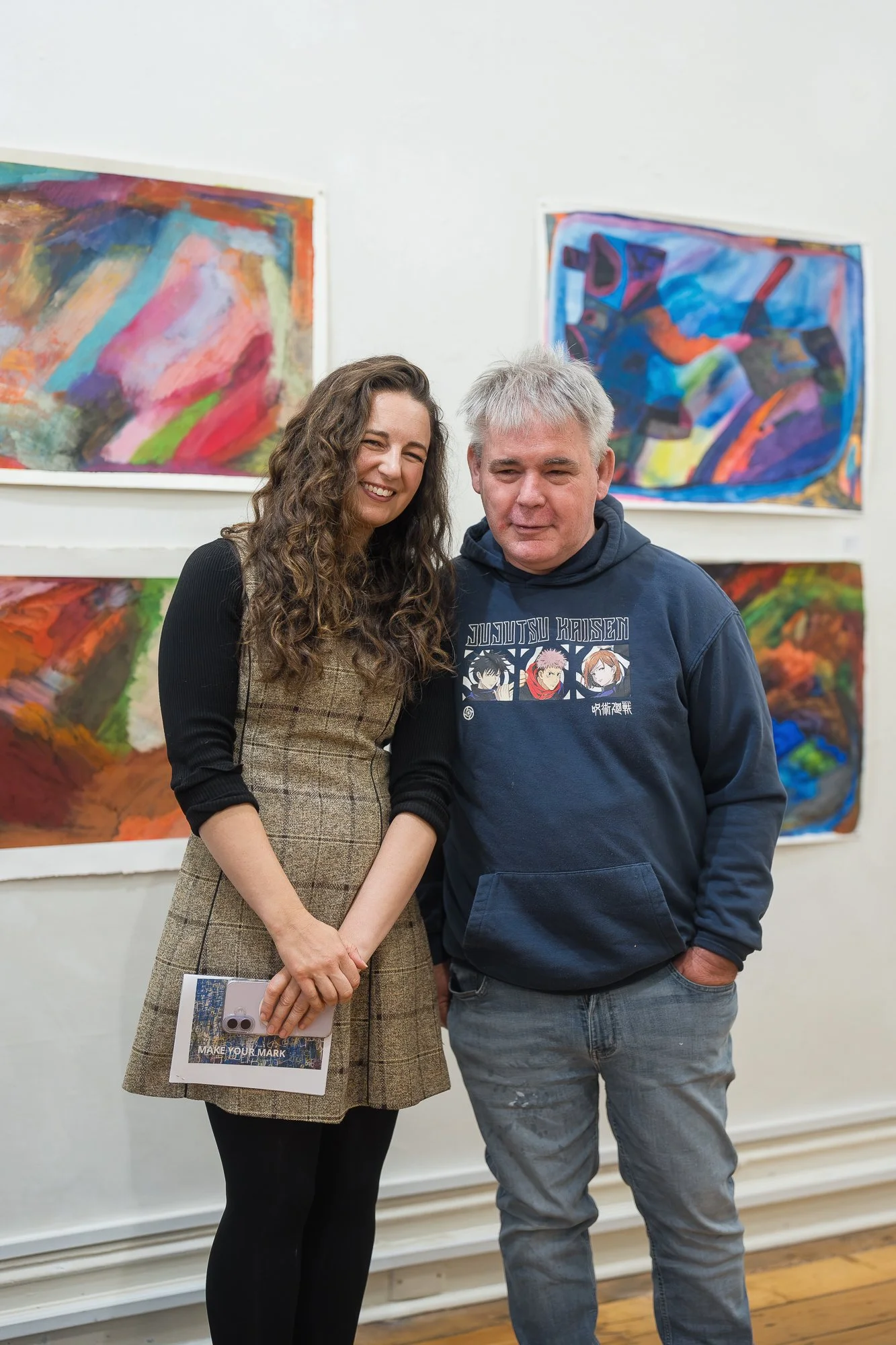 Two people smile together in front of a wall of bold, expressive works on paper — one holding a Make Your Mark catalogue.