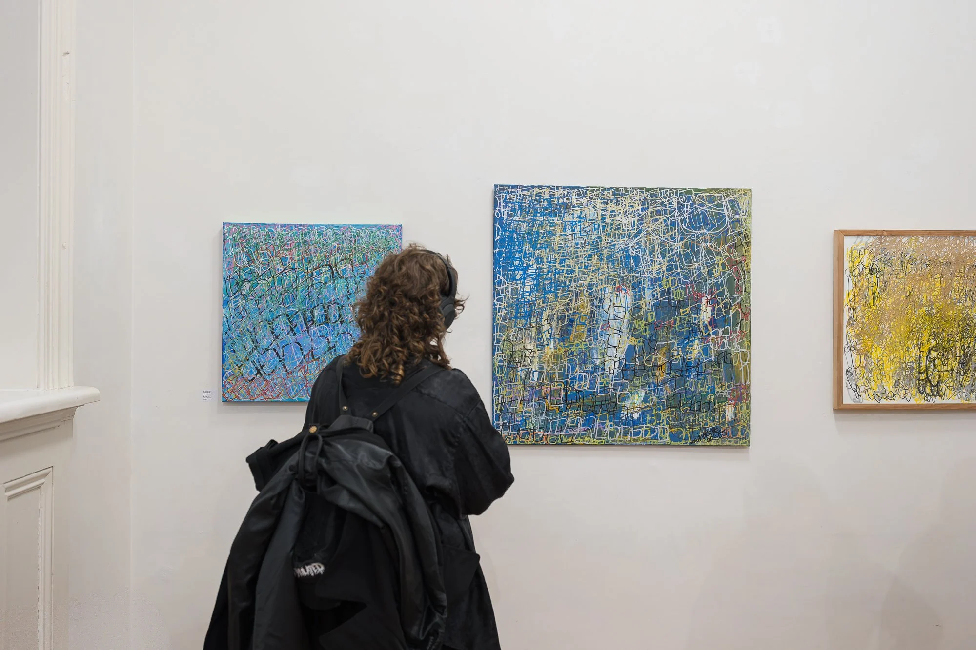 A visitor in a black coat stands before a series of densely layered abstract paintings in blue and green.