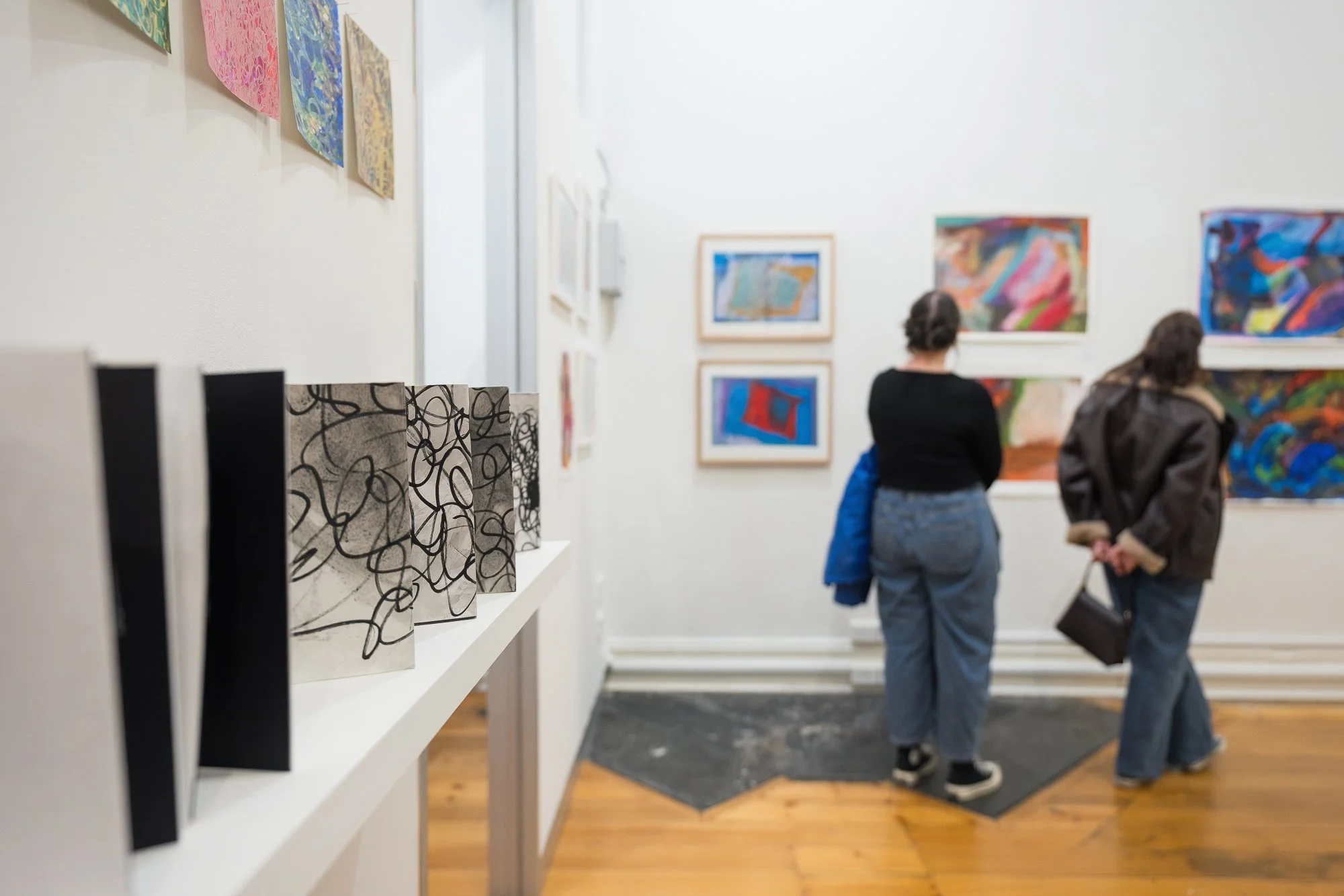 Black-and-white works on a plinth in the foreground, with two visitors taking in a wall of vivid colourful paintings beyond.