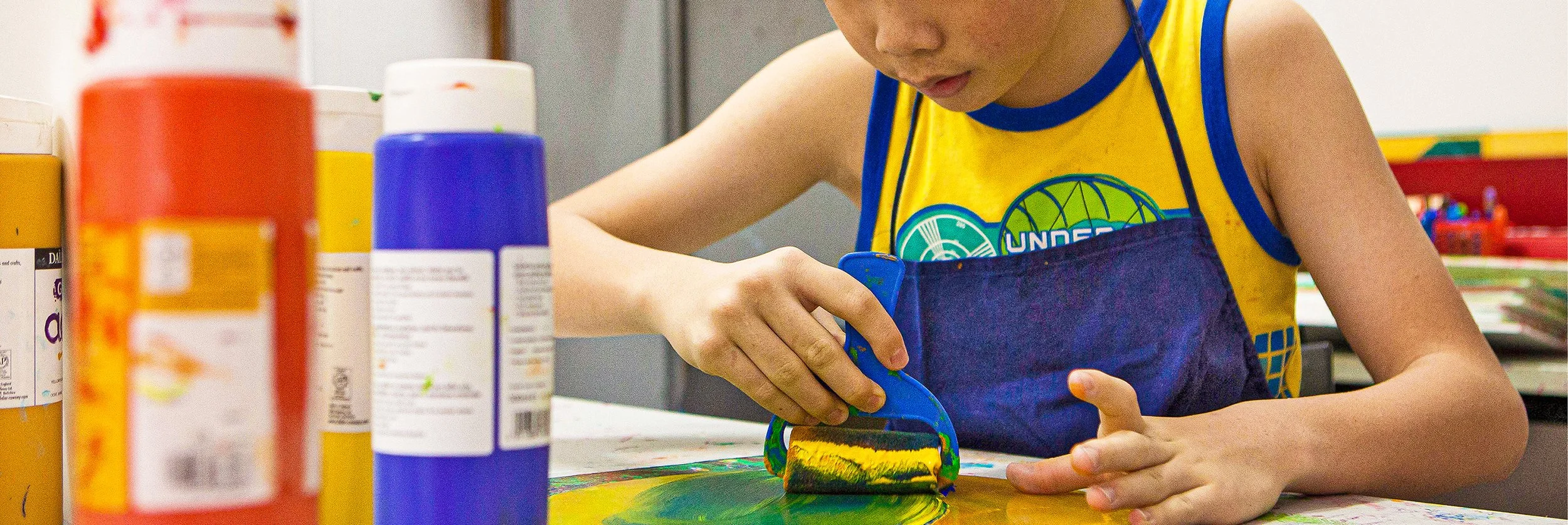 A close up of an artist in a blue apron from a supported studio working on a blue, yellow and green print or painting.