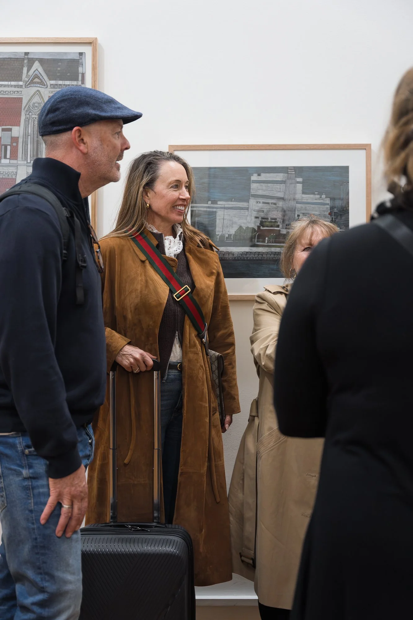 Group of four visitors in conversation near framed architectural illustrations, woman with a rolling suitcase.