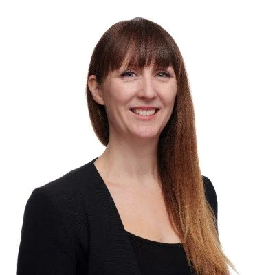 Woman with long brown hair and bangs, wearing a black top, smiling against a white background.