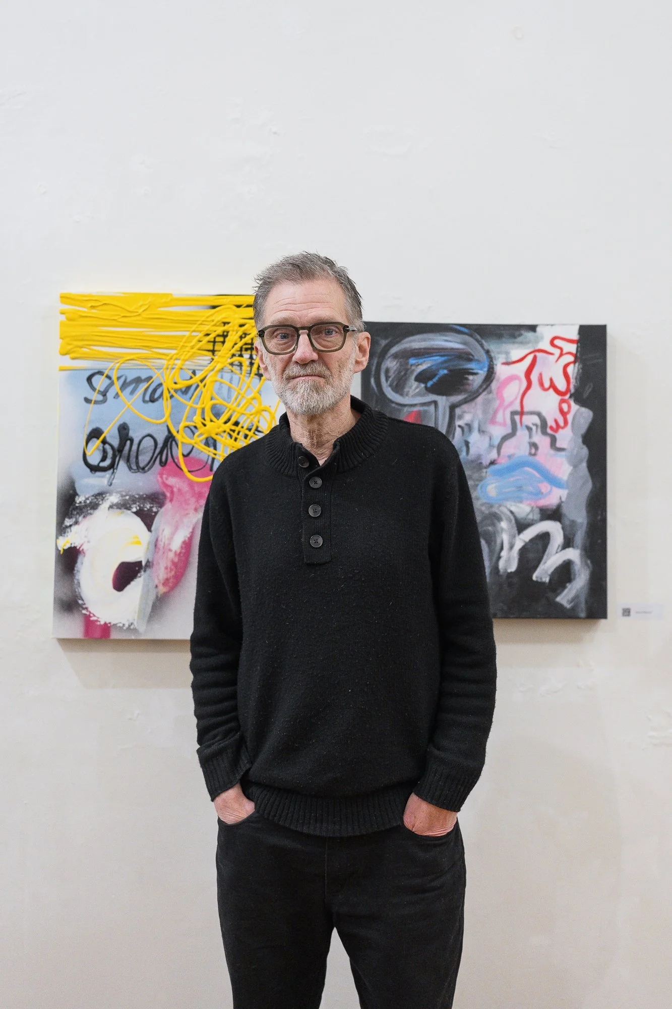 Portrait of a bearded man in black jumper standing in front of abstract mixed-media paintings with handwritten text ("Rot," "Tremor").
