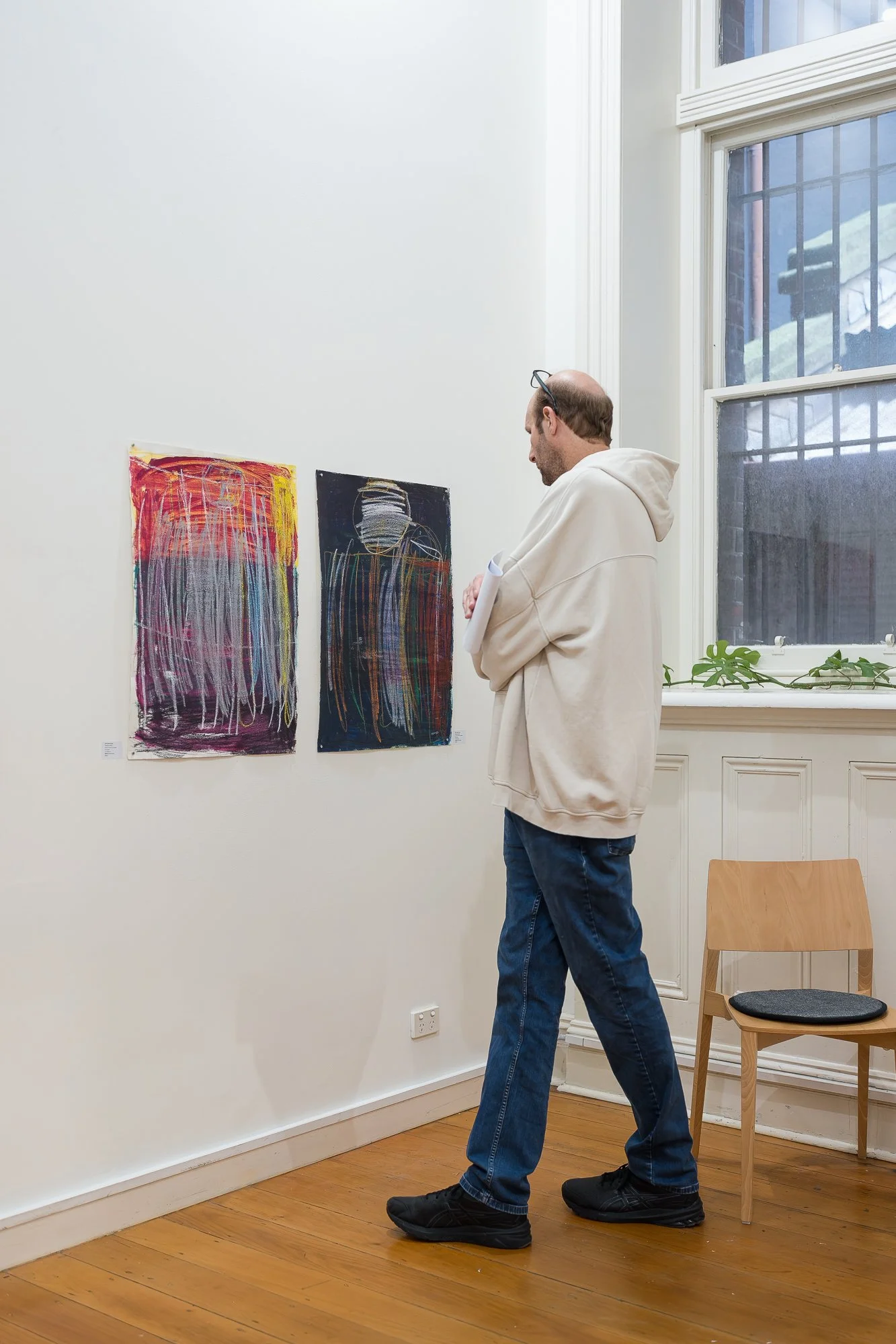 A visitor in a cream hoodie moves slowly along the wall, taking time with two expressive works on paper in red, yellow and dark blue.