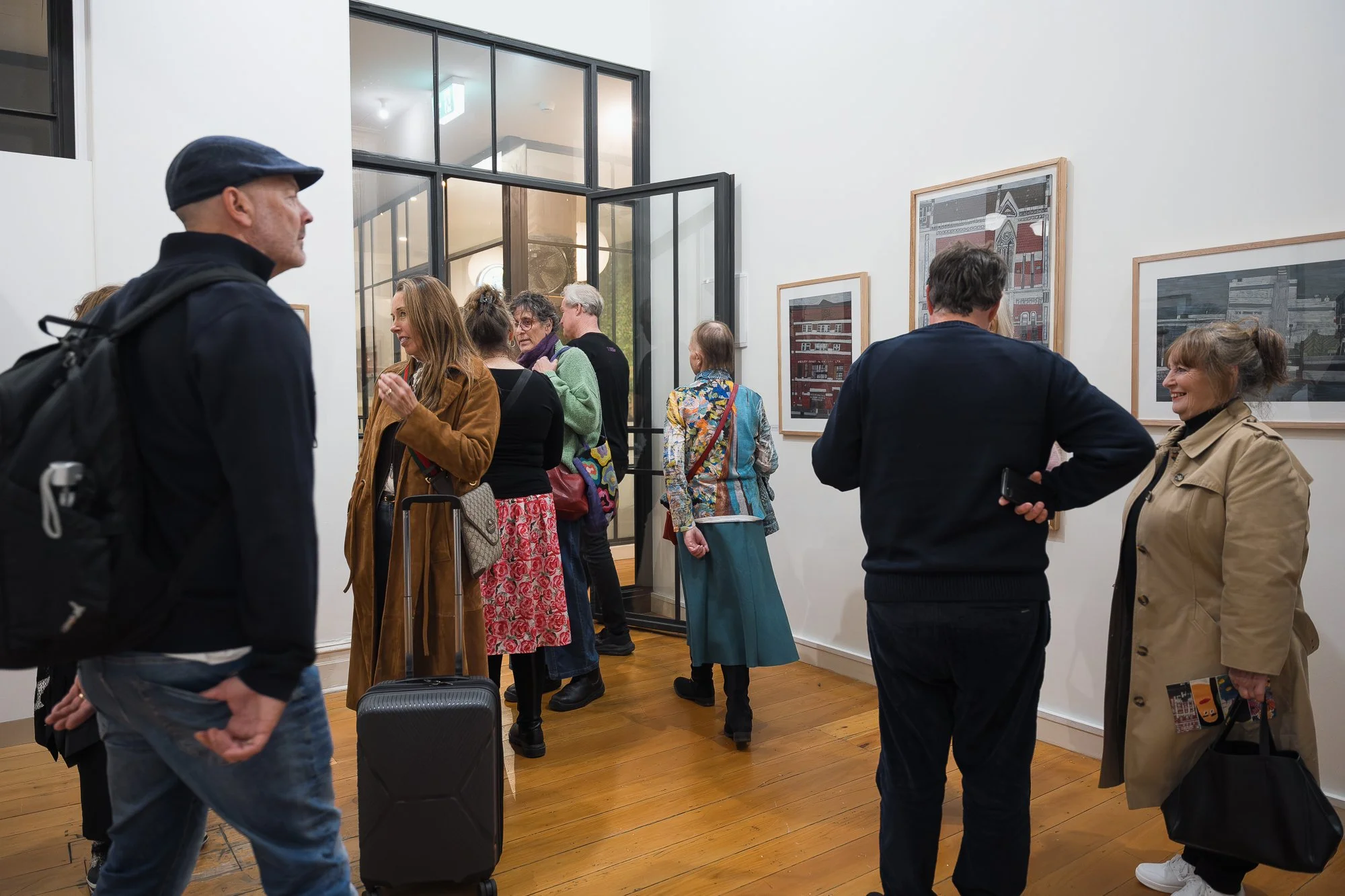 Group of four visitors examining a framed architectural illustration, woman in patterned jacket with rolling suitcase nearby.