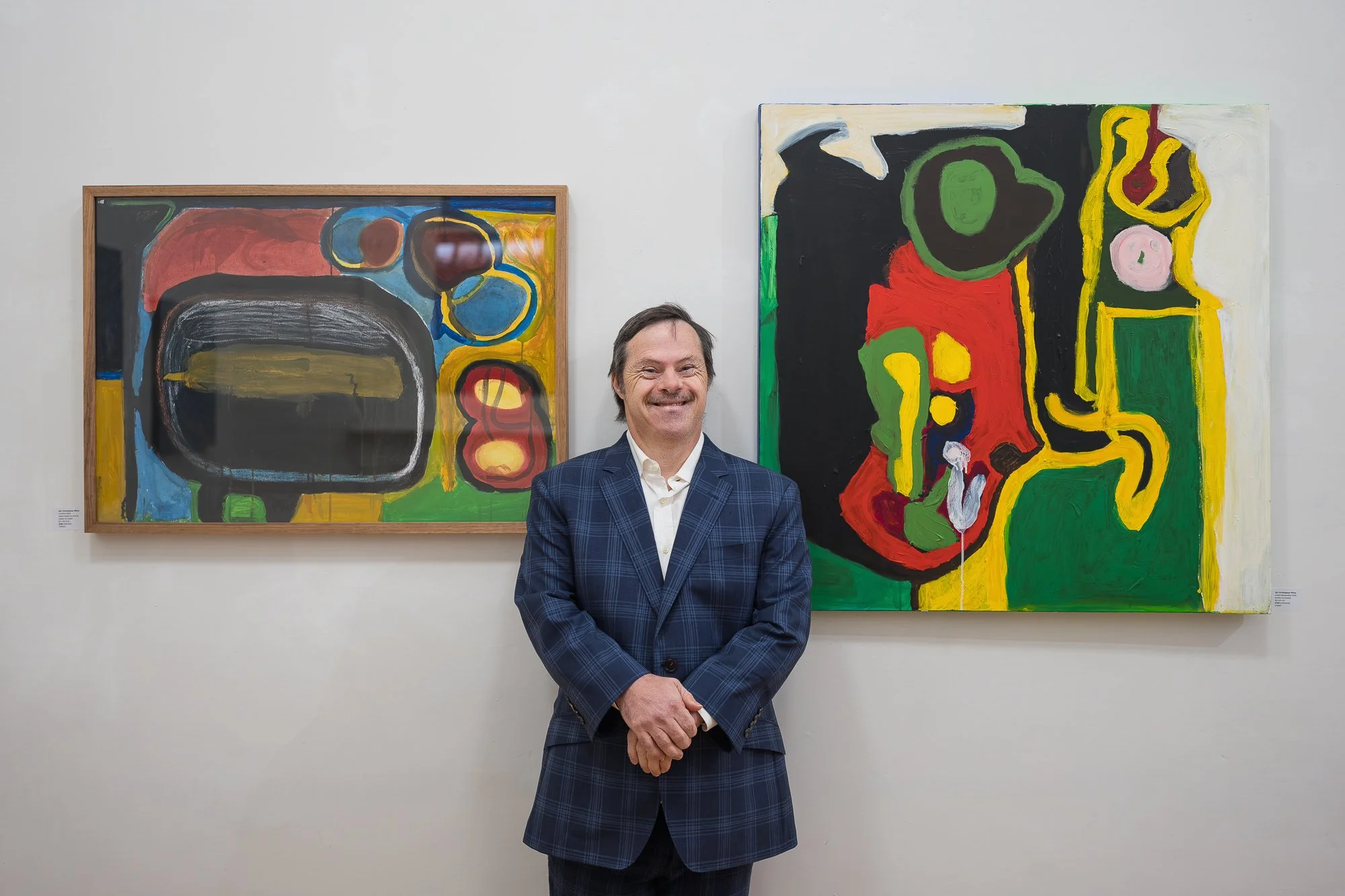 A smiling man in a navy check blazer stands in front of two large, bold abstract paintings.