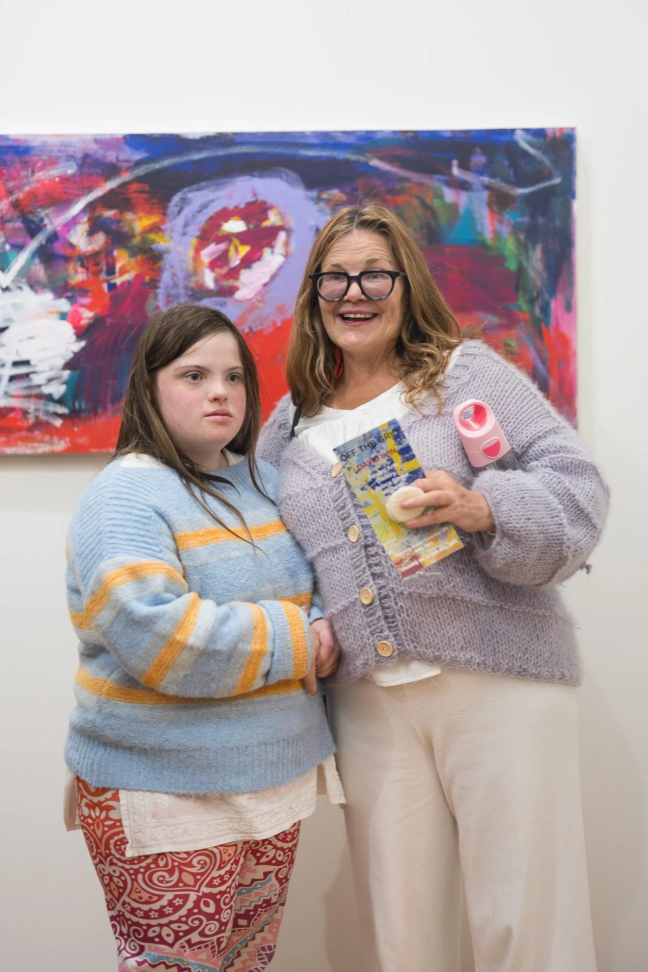 Two people smile together in front of a large, vivid abstract painting in rich reds and blues.