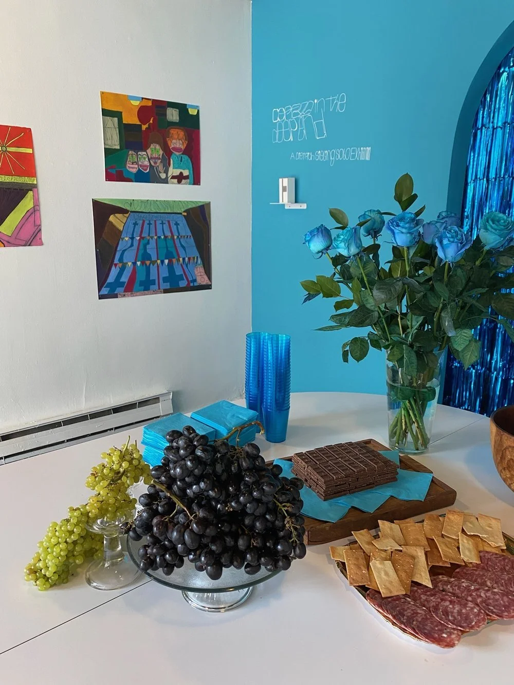 Buffet table with grapes, blue roses, salami, and crackers, in a room with colorful paintings and blue decor.