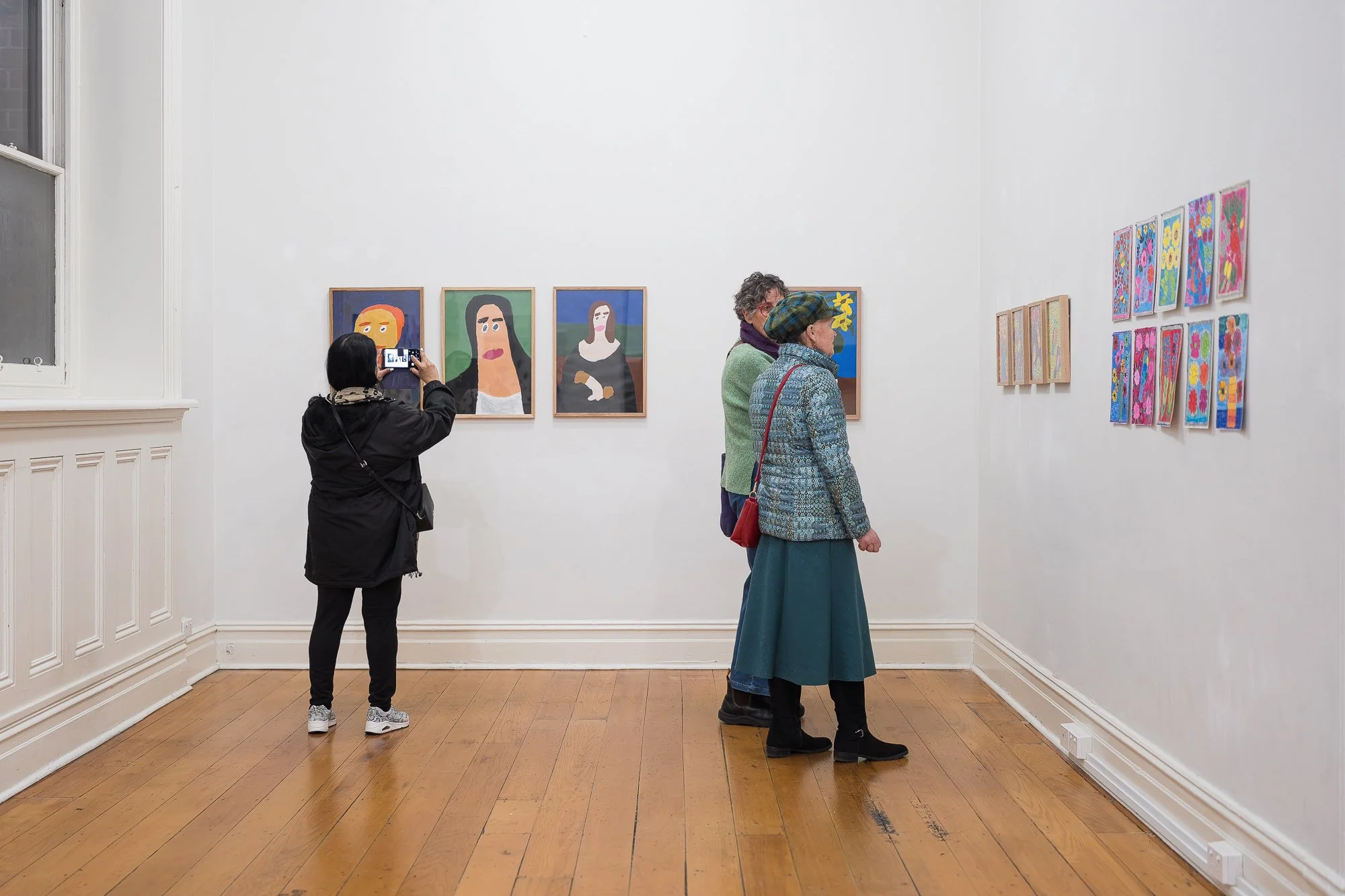Visitor photographing a row of three portrait paintings on a gallery wall; two other visitors viewed from behind further along the same wall, with smaller framed works visible on the right-hand wall.