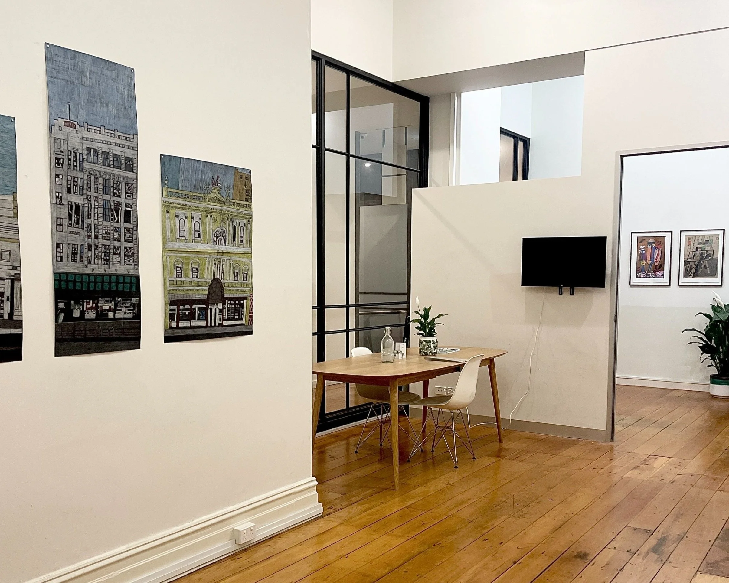 Project space interior with wooden floors, a table with chairs, wall art depicting buildings, and a TV mounted on the wall.