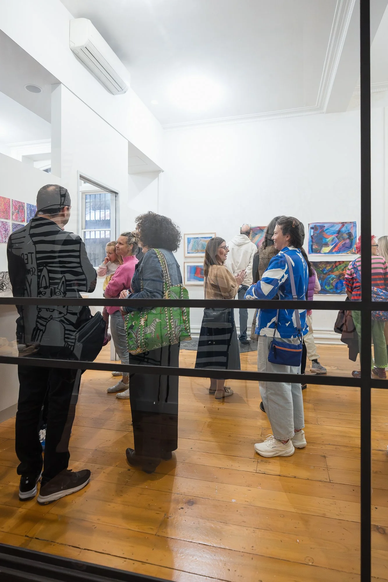 A lively crowd fills the main gallery space, viewed through the glass partition, works covering every wall.