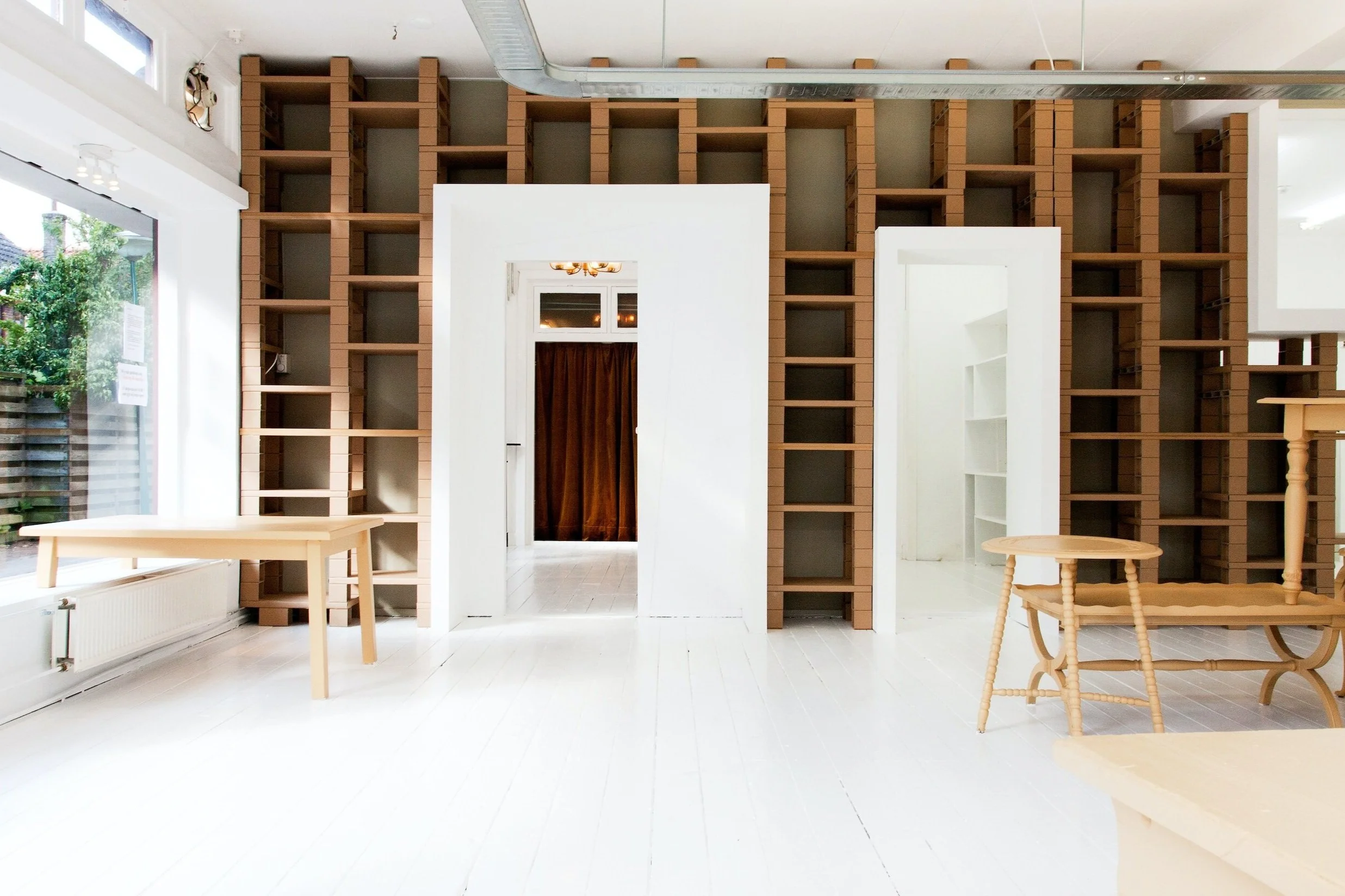 Light filled white room, with large window and empty wooden tables and floor to ceiling bookshelves.