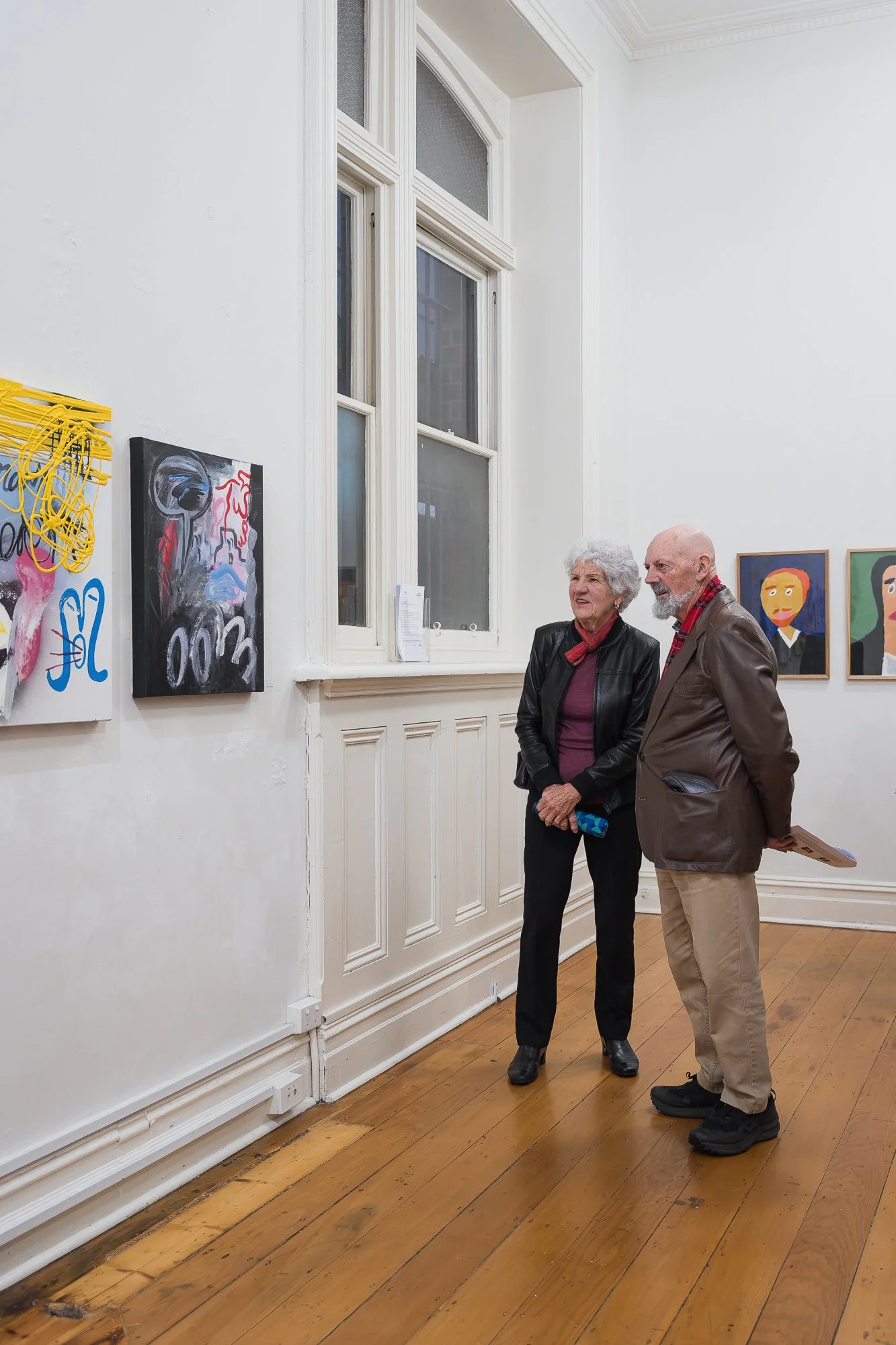 Older couple standing together viewing an abstract painting and portrait works on the gallery wall, near a tall window with an event flyer on the sill.